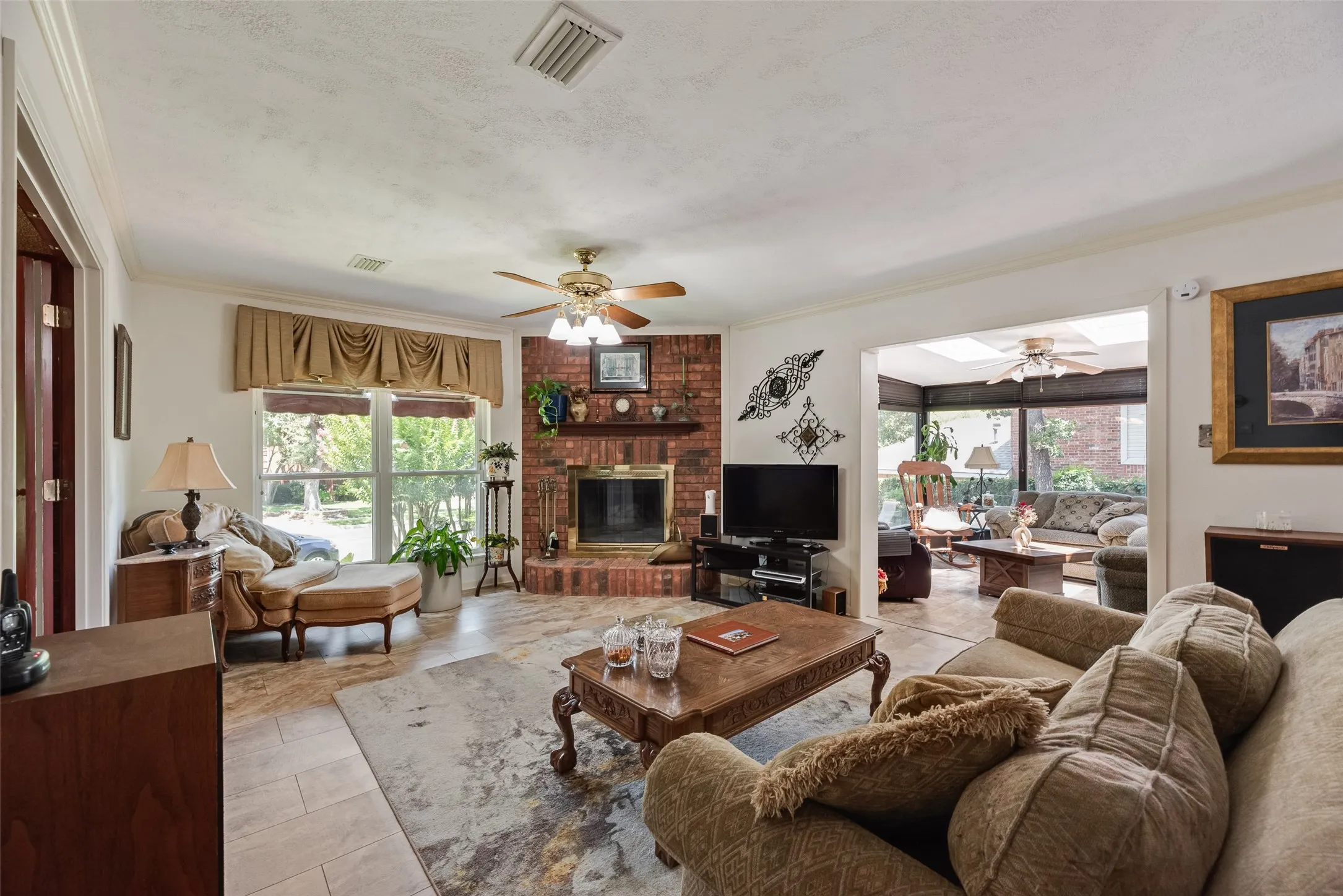 Living area featuring a brick fireplace, ceiling fan with integrated lighting, tiled flooring, and a large window overlooking exterior landscaping