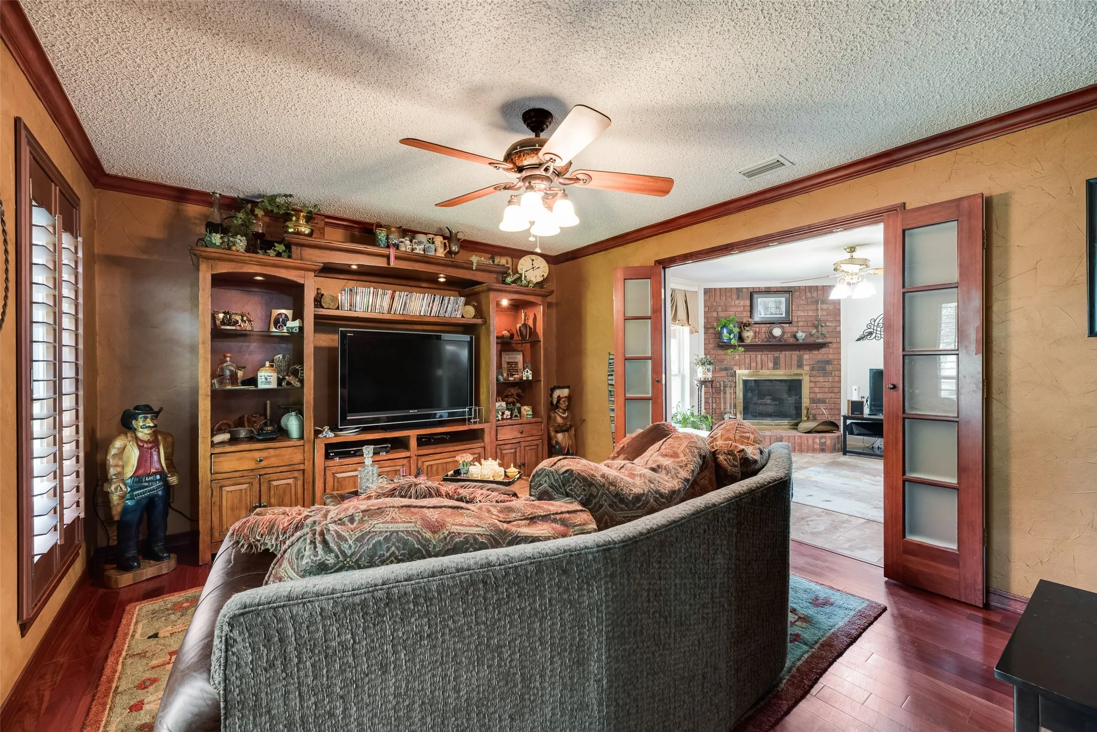 Wood-finish flooring, built-in entertainment center with shelving, ceiling fan, and double wood-frame doors with frosted glass panels