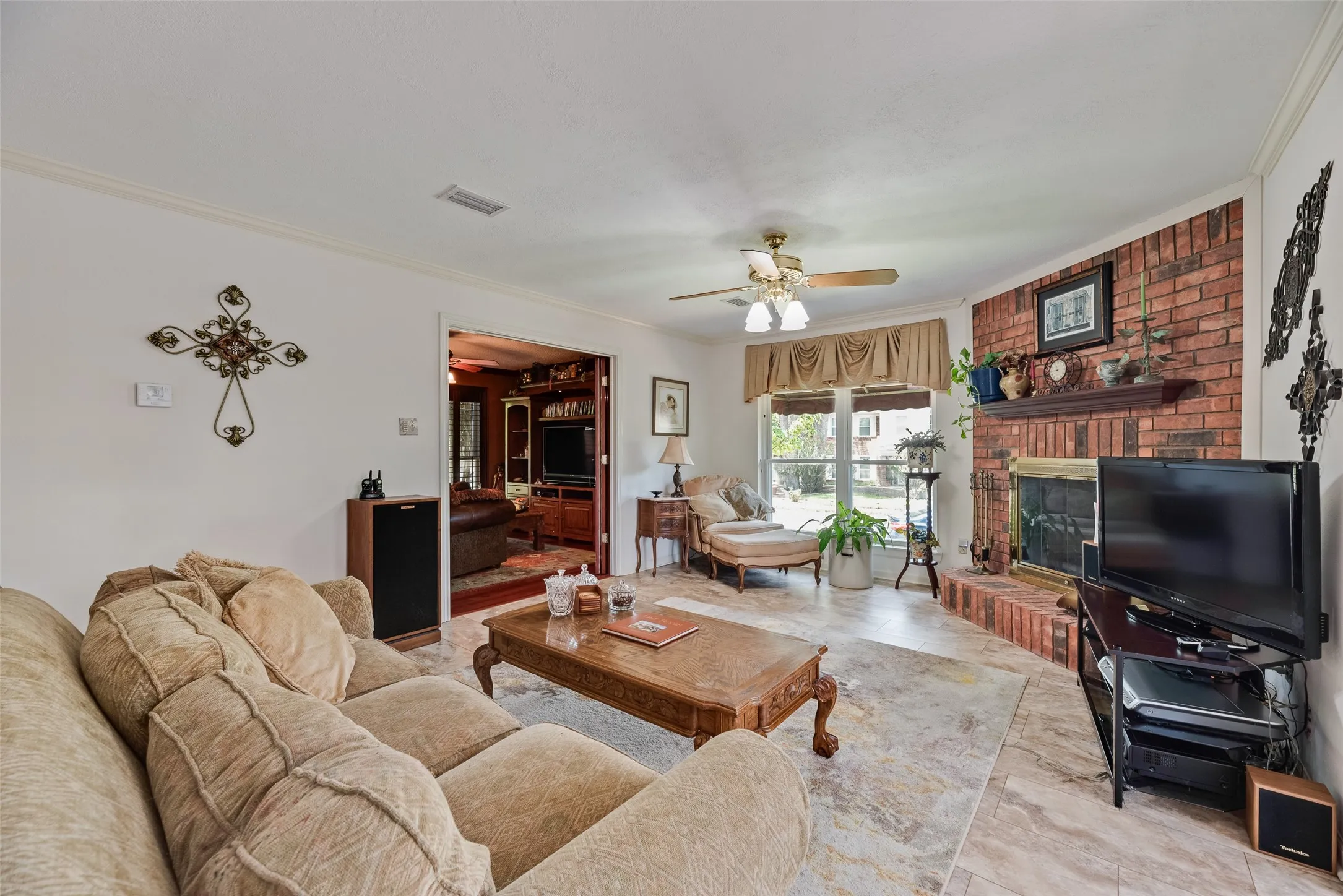 Living area with tile flooring, a brick fireplace with a wood mantle, a ceiling fan with integrated lighting, and a large window