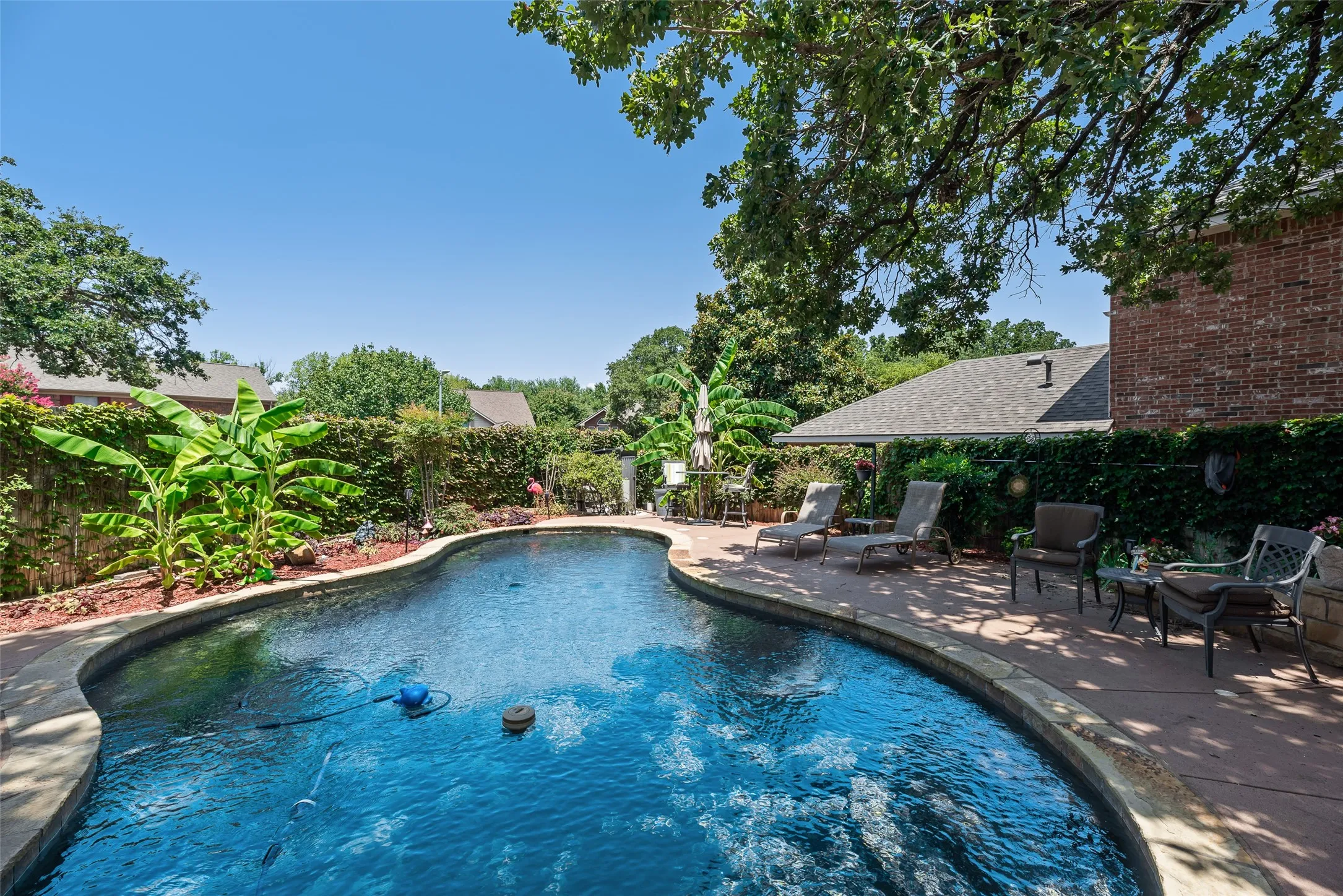Freeform swimming pool with a surrounding concrete patio, mature landscaping, and a brick exterior structure