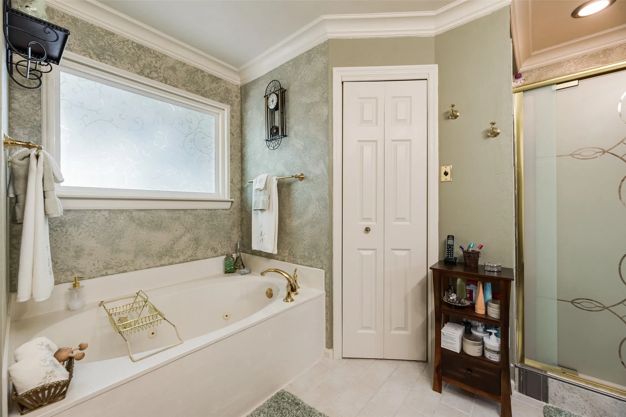 Jetted bathtub with brass fixtures, frosted window, tiled flooring, and a glass-enclosed shower with decorative etching
