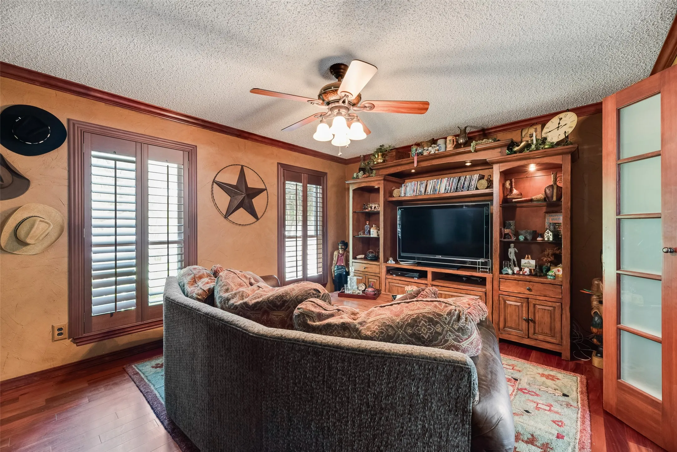 Wood-finish flooring, built-in entertainment center, ceiling fan, crown molding, and two windows with plantation shutters