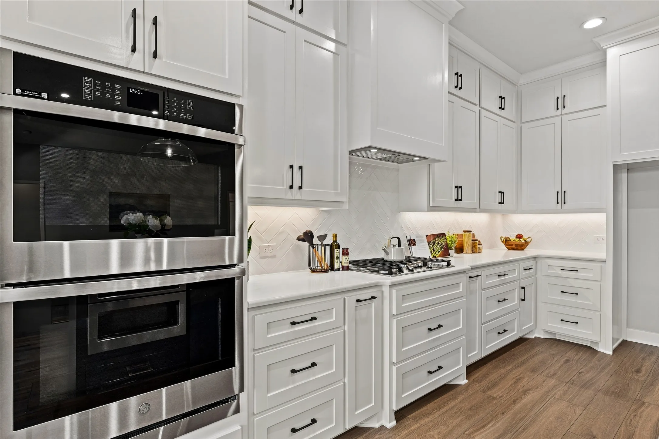 White cabinetry with matte black hardware, stainless steel built-in appliances, herringbone tile backsplash, wood-finish flooring, and under-cabinet lighting