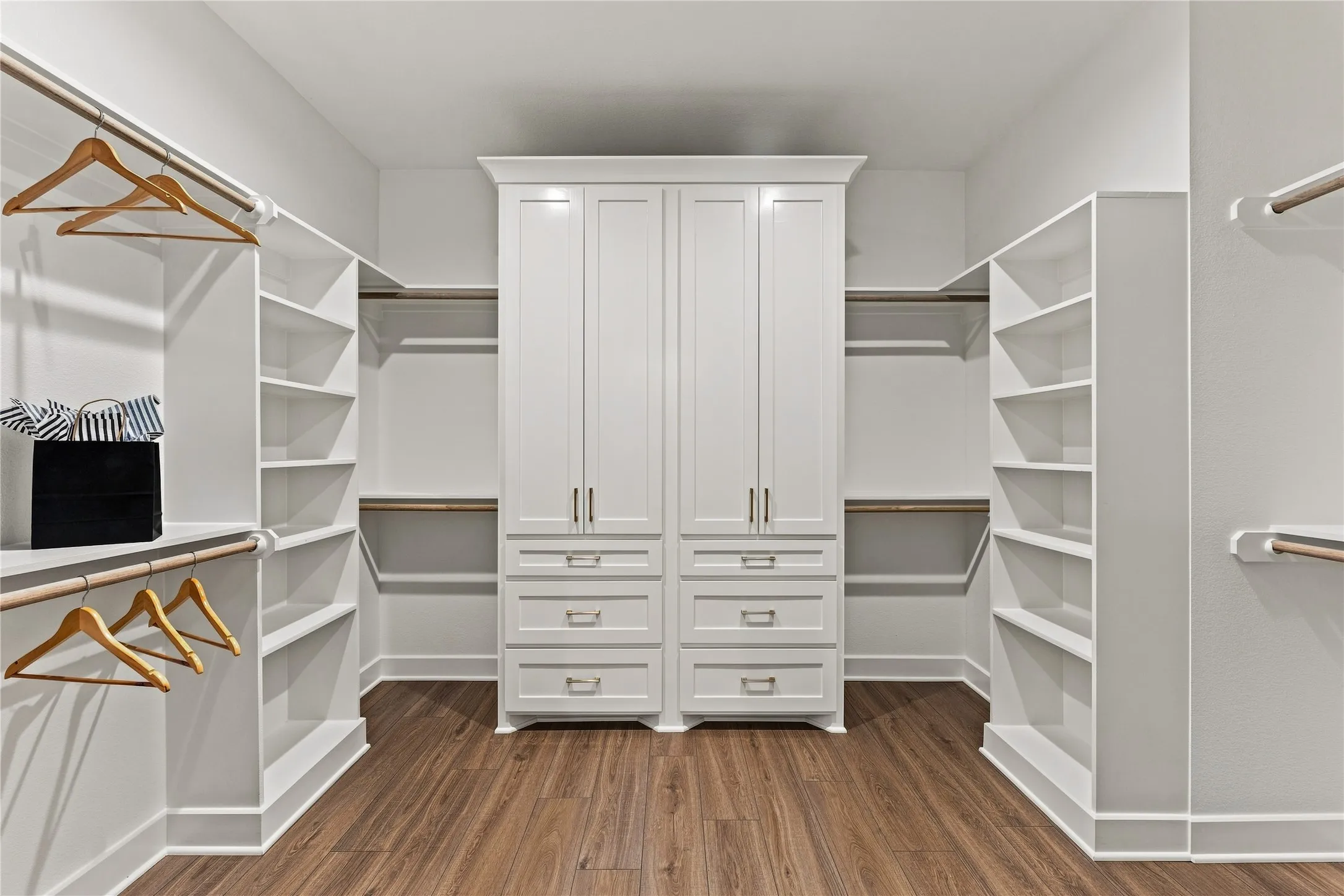 Walk-in closet featuring wood-finish flooring, built-in shelving, hanging rods, and a central cabinet with drawers and closed storage