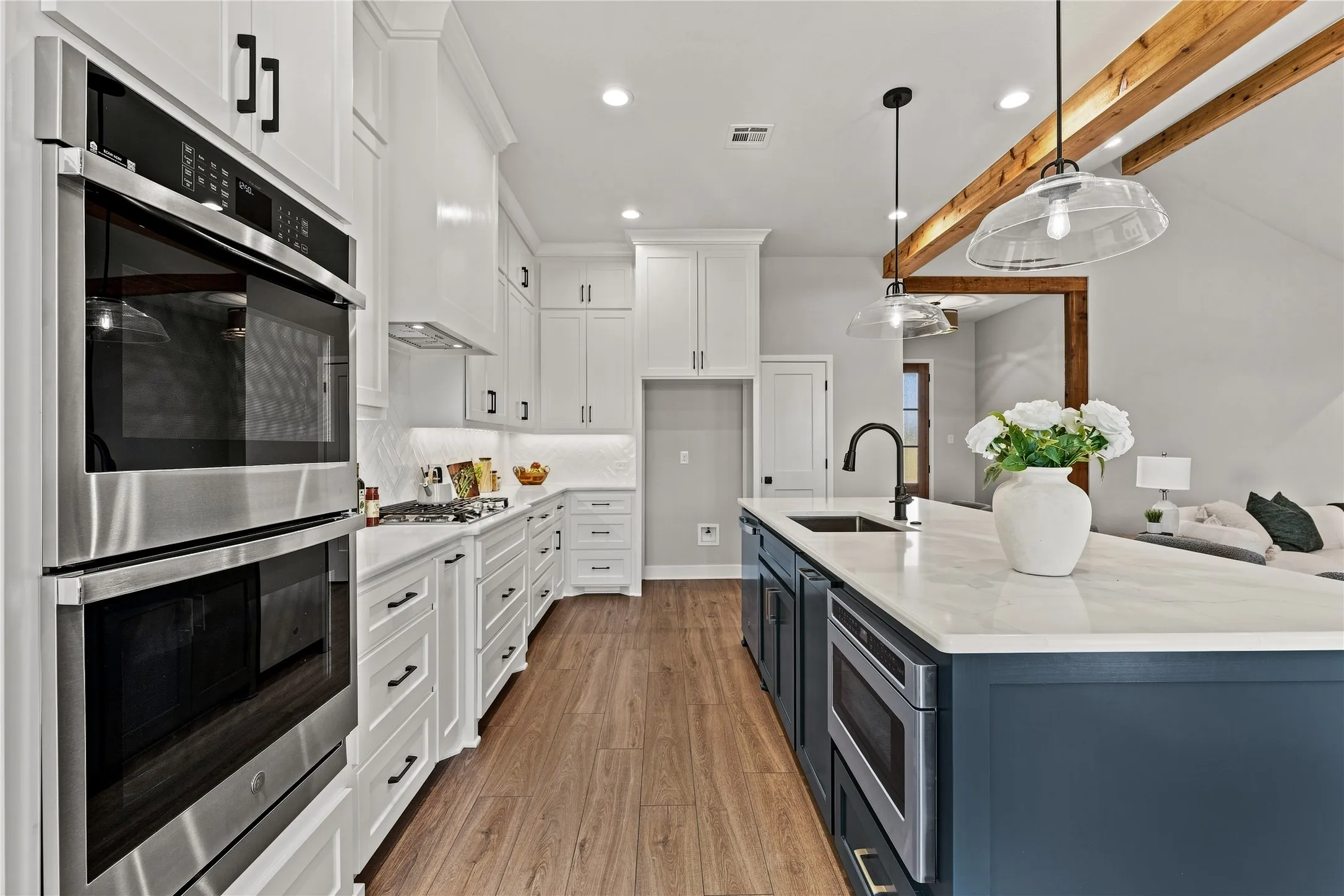 White cabinetry with matte black hardware, stainless steel double wall ovens, wood-finish flooring, a kitchen island with integrated microwave, and exposed wood ceiling beams