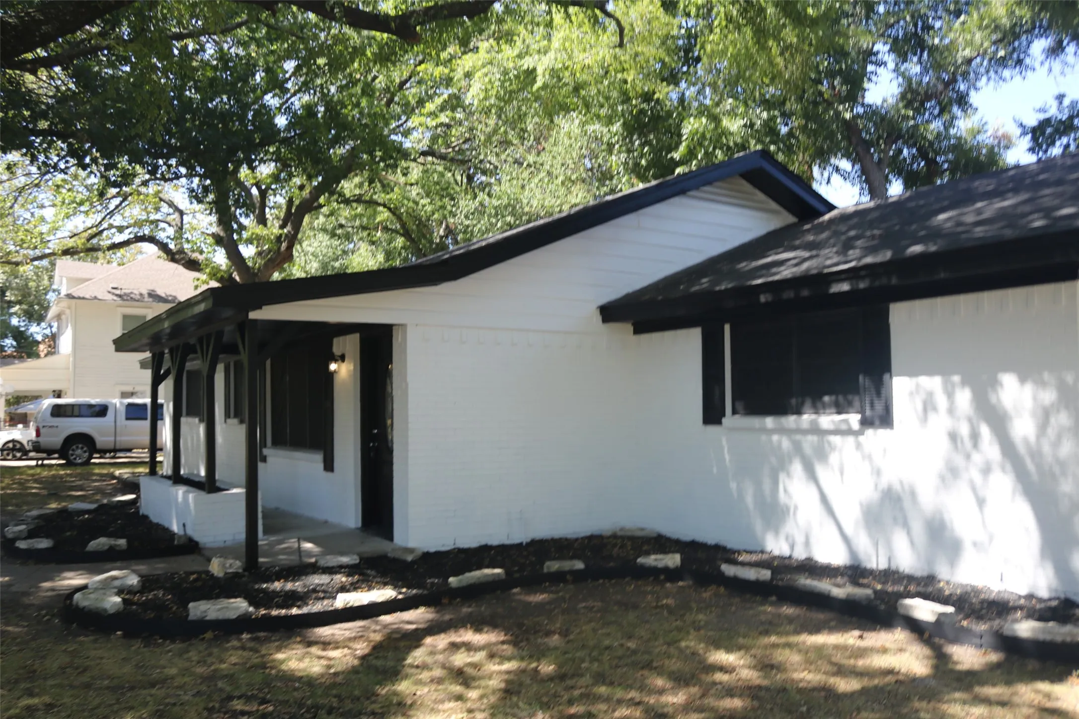 White exterior with a covered porch featuring dark posts, black-trimmed windows, a dark shingle roof, and landscaped garden beds