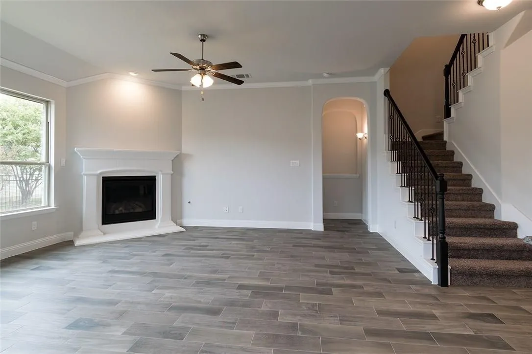 Living area with wood-finish flooring, corner fireplace with white surround, ceiling fan, arched entryway, and staircase with wrought iron balusters
