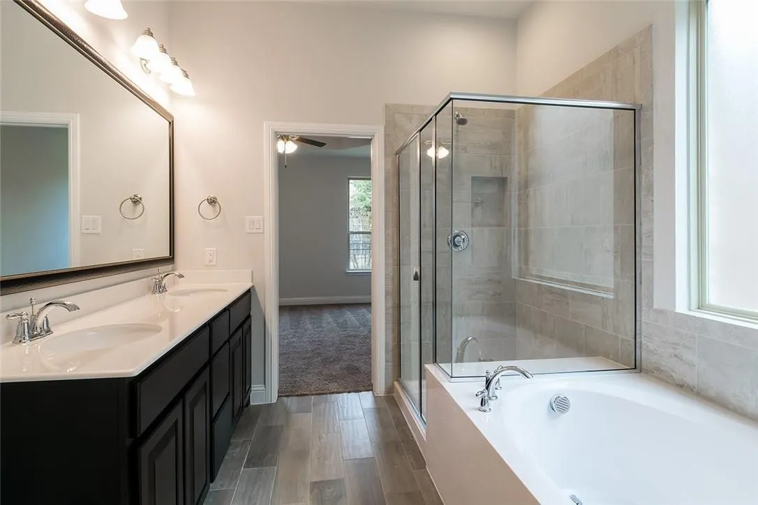 Dual vanity with undermount sinks, wood-finish flooring, tiled shower with glass enclosure, built-in bathtub, and privacy window