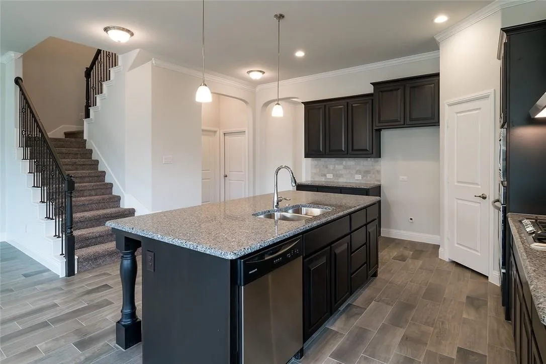 Spacious kitchen island with a double basin sink and granite countertop, dark wood-finish cabinetry, stainless steel dishwasher, and wood-look tile flooring