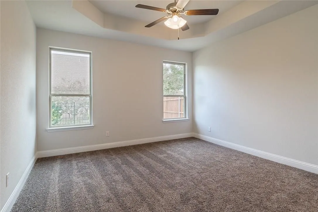 Tray ceiling with a ceiling fan, two windows, and neutral carpeting