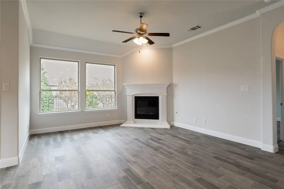 Wood-finish flooring, two large windows, an electric fireplace with a white mantel, a ceiling fan, and crown molding