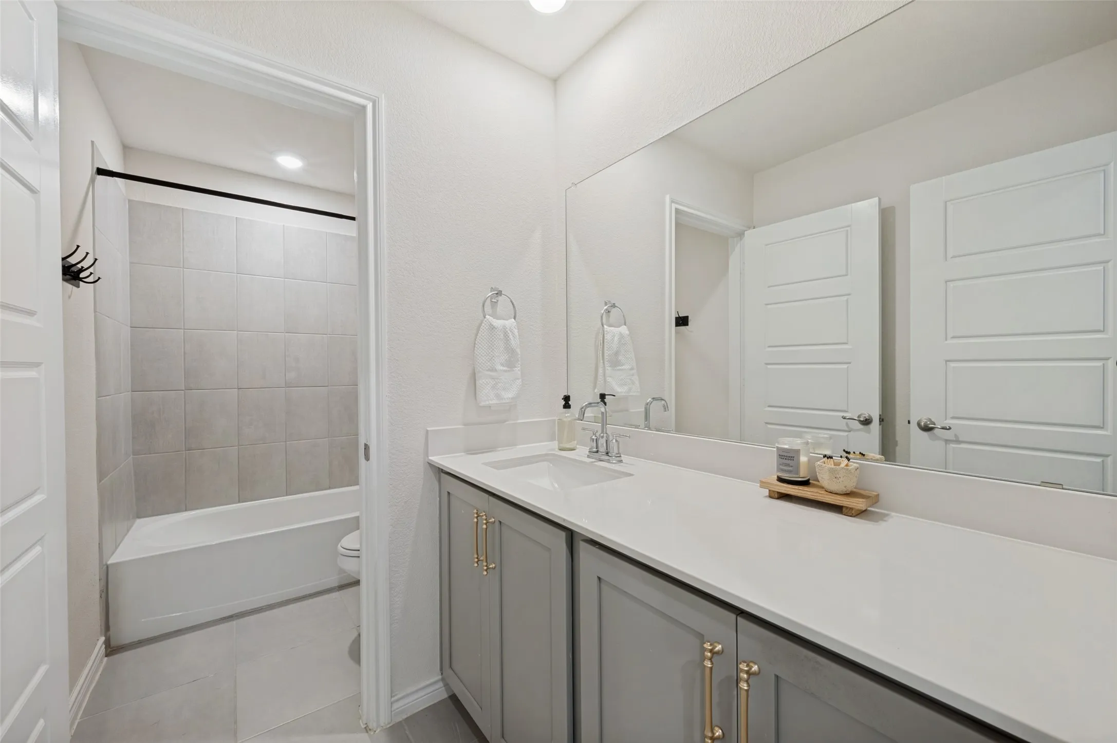 Full-width vanity mirror, gray shaker-style cabinetry with gold-tone hardware, white countertop, undermount sink, and tiled tub surround