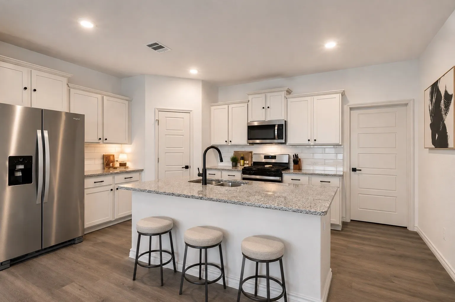 Modern kitchen featuring a large island with granite-finish countertop, white cabinetry with black hardware, stainless steel appliances, and wood-finish flooring