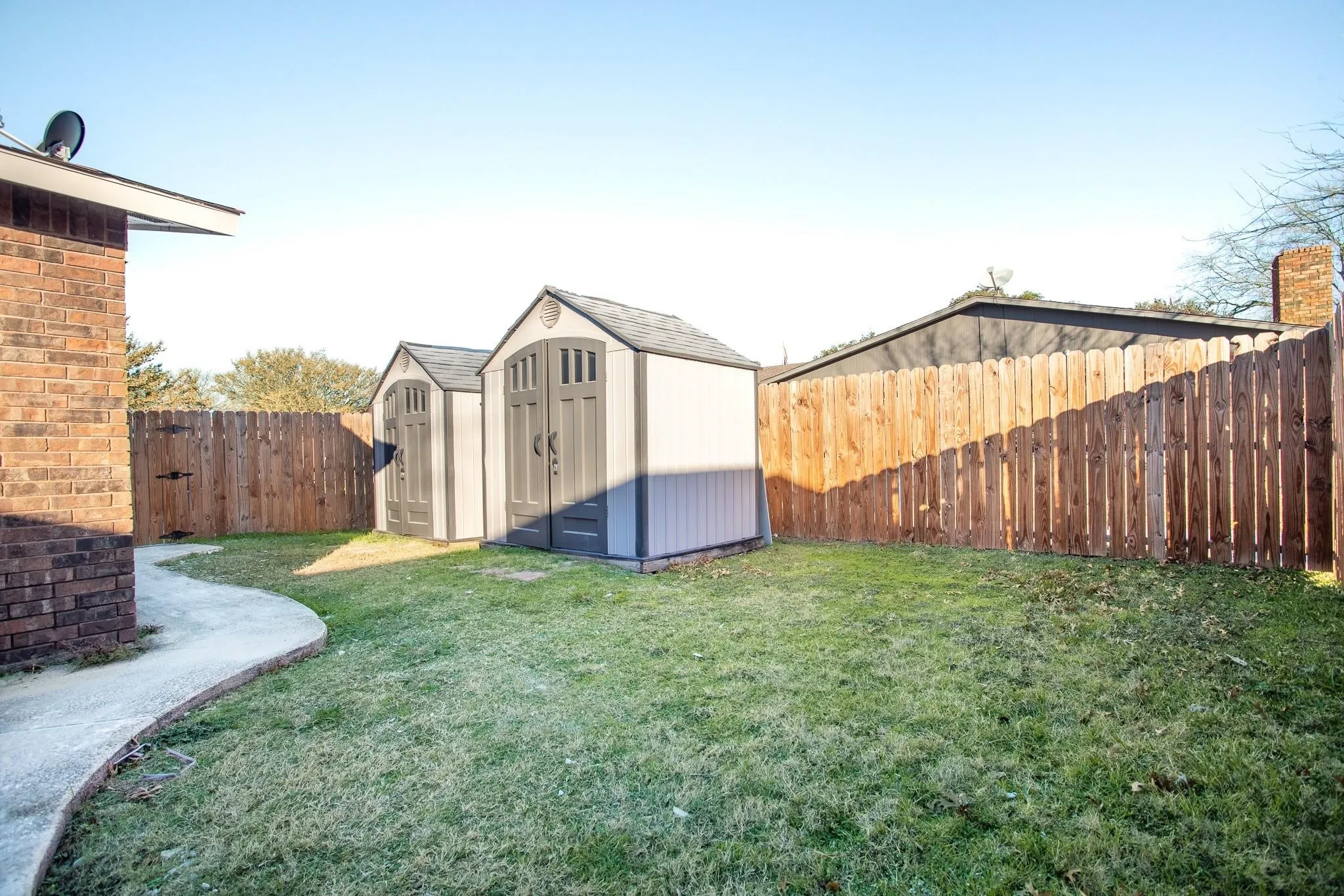 Expansive backyard featuring a manicured lawn, two storage sheds, a privacy fence, and a curved concrete pathway
