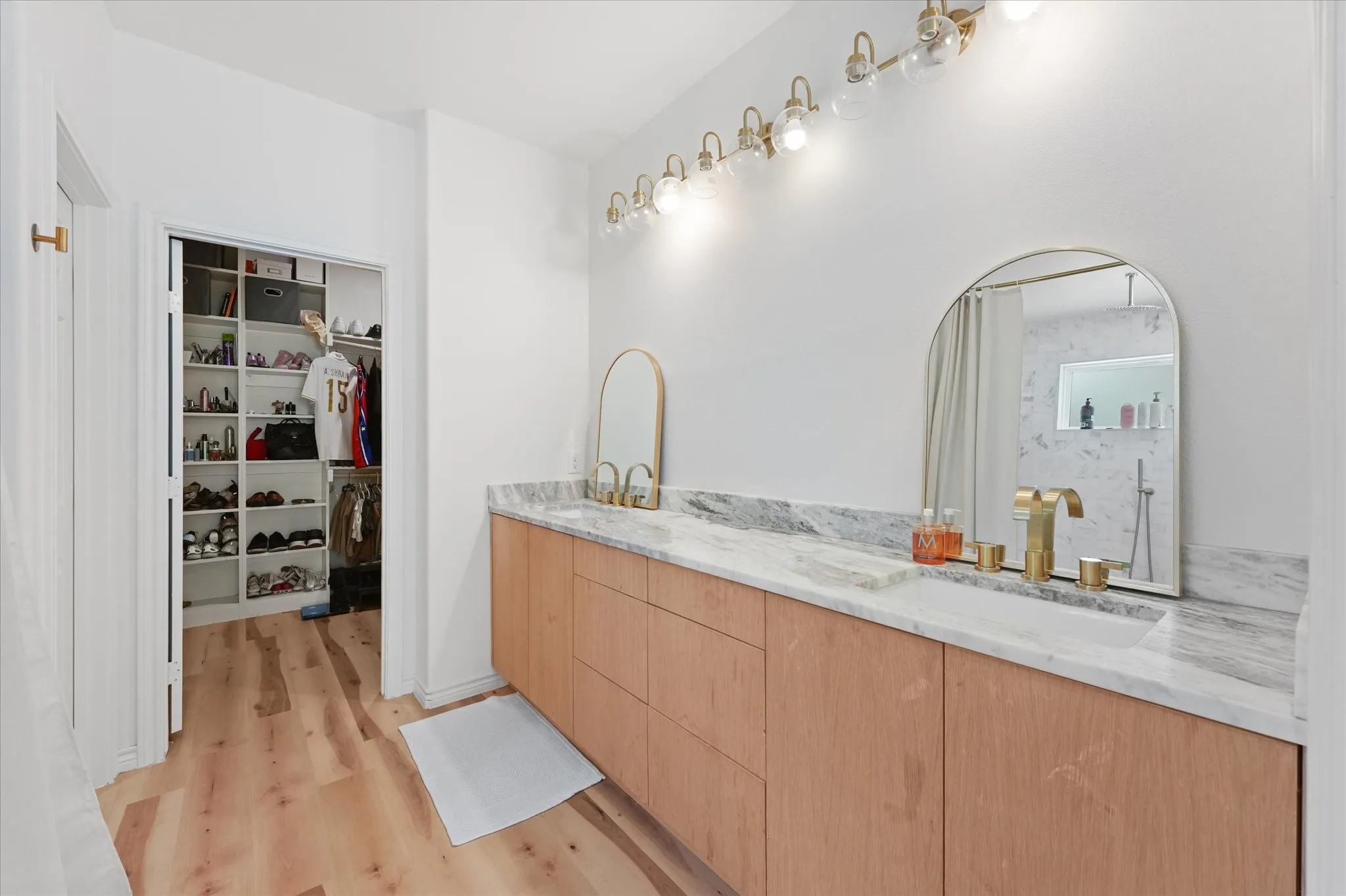Light wood-finish flooring, dual vanity with light wood cabinetry, stone-finish countertop, and gold-tone fixtures