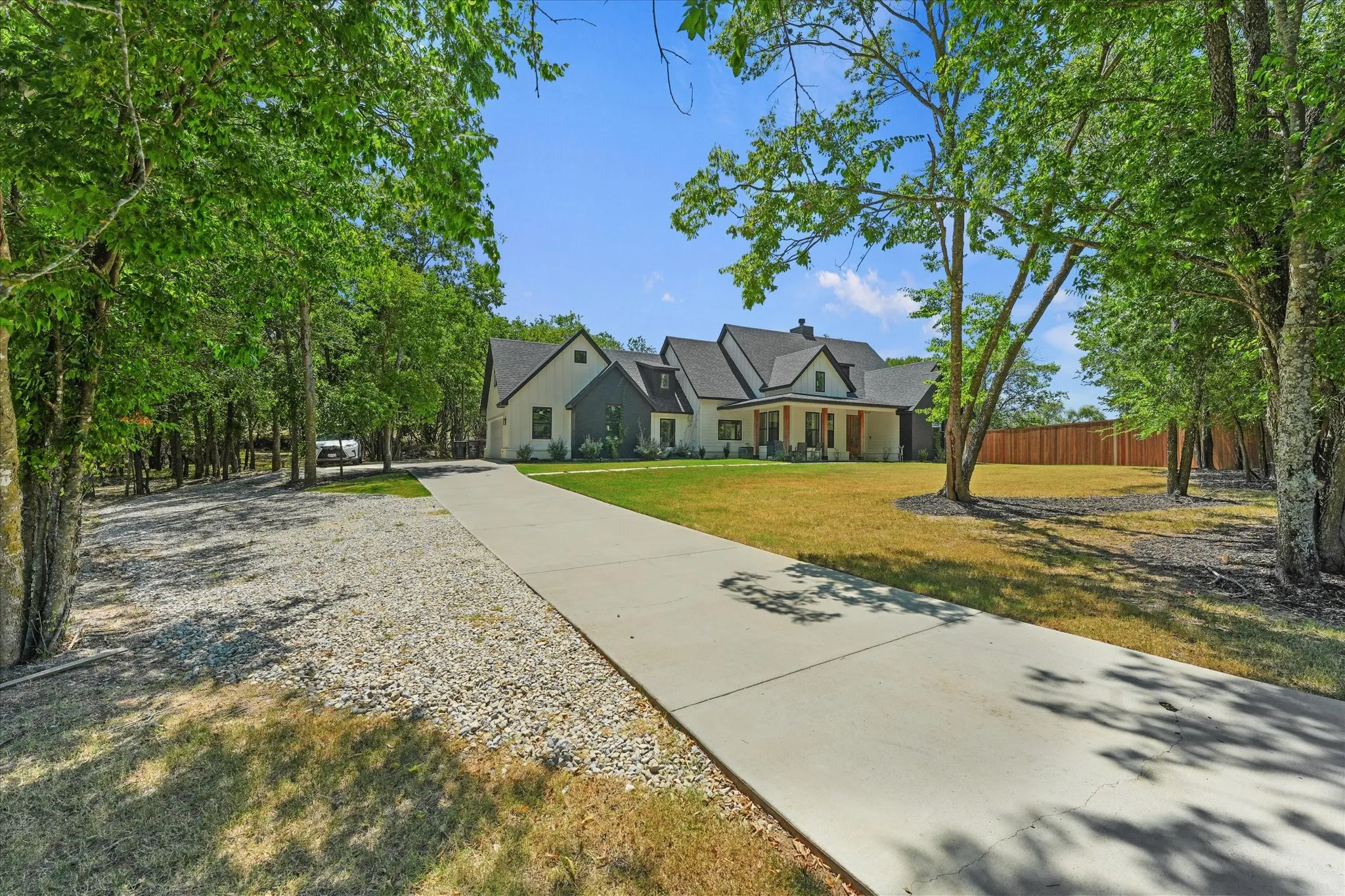 Modern farmhouse exterior with a gabled roof, a concrete driveway, a gravel parking area, and a wood privacy fence