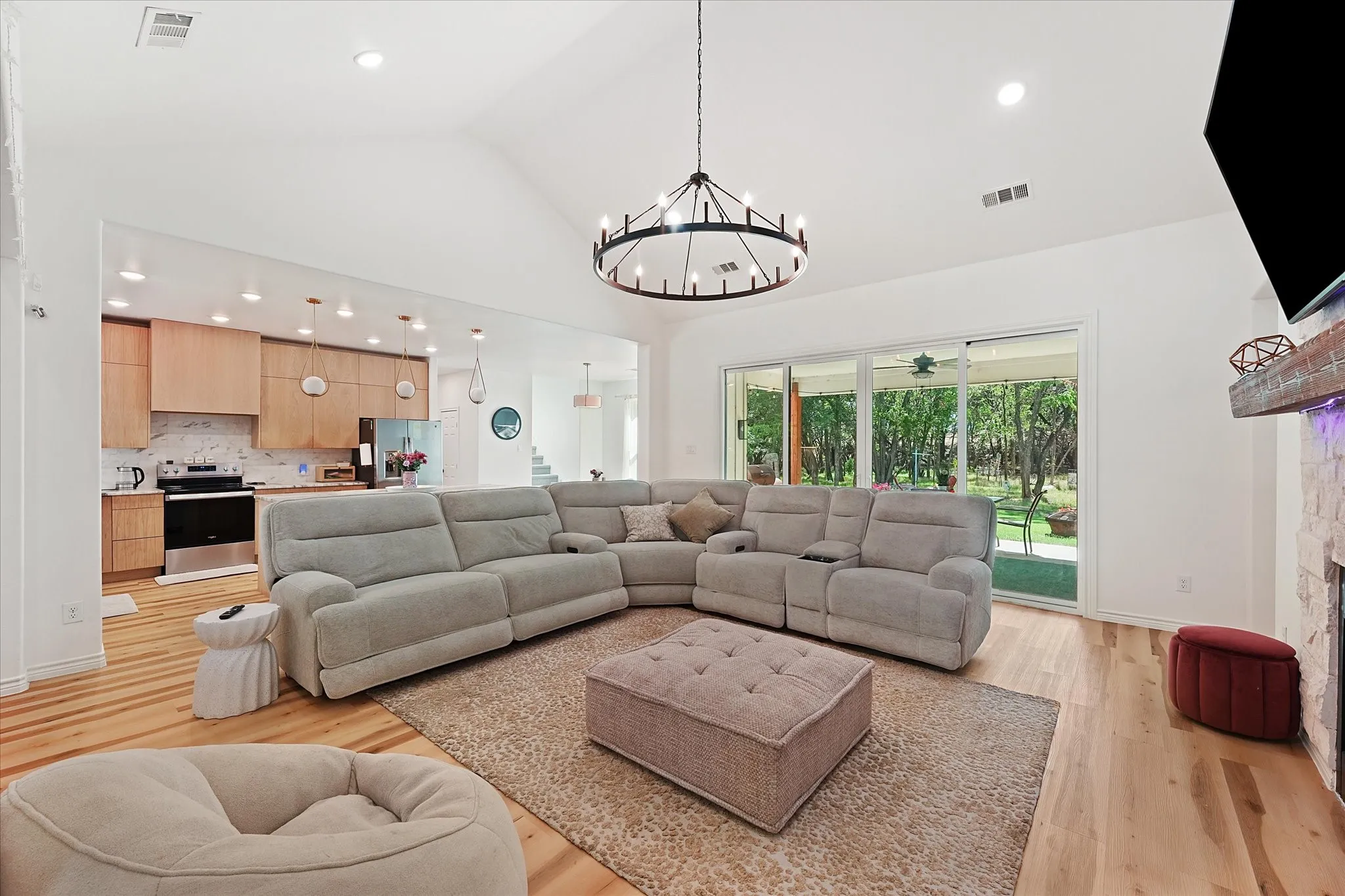 Vaulted ceilings with a chandelier, wood-finish flooring, a stone fireplace, sliding glass doors, and a kitchen with light wood cabinetry