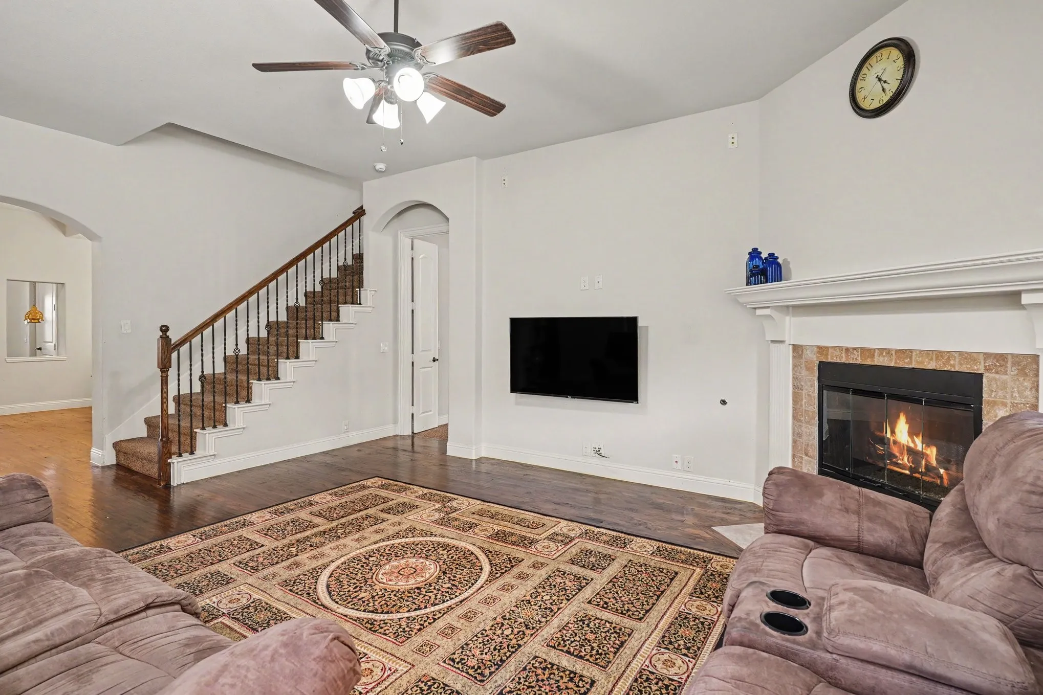 Fireplace with tile surround and mantel, wood-finish flooring, staircase with wrought iron balusters, ceiling fan with light fixture, arched doorways