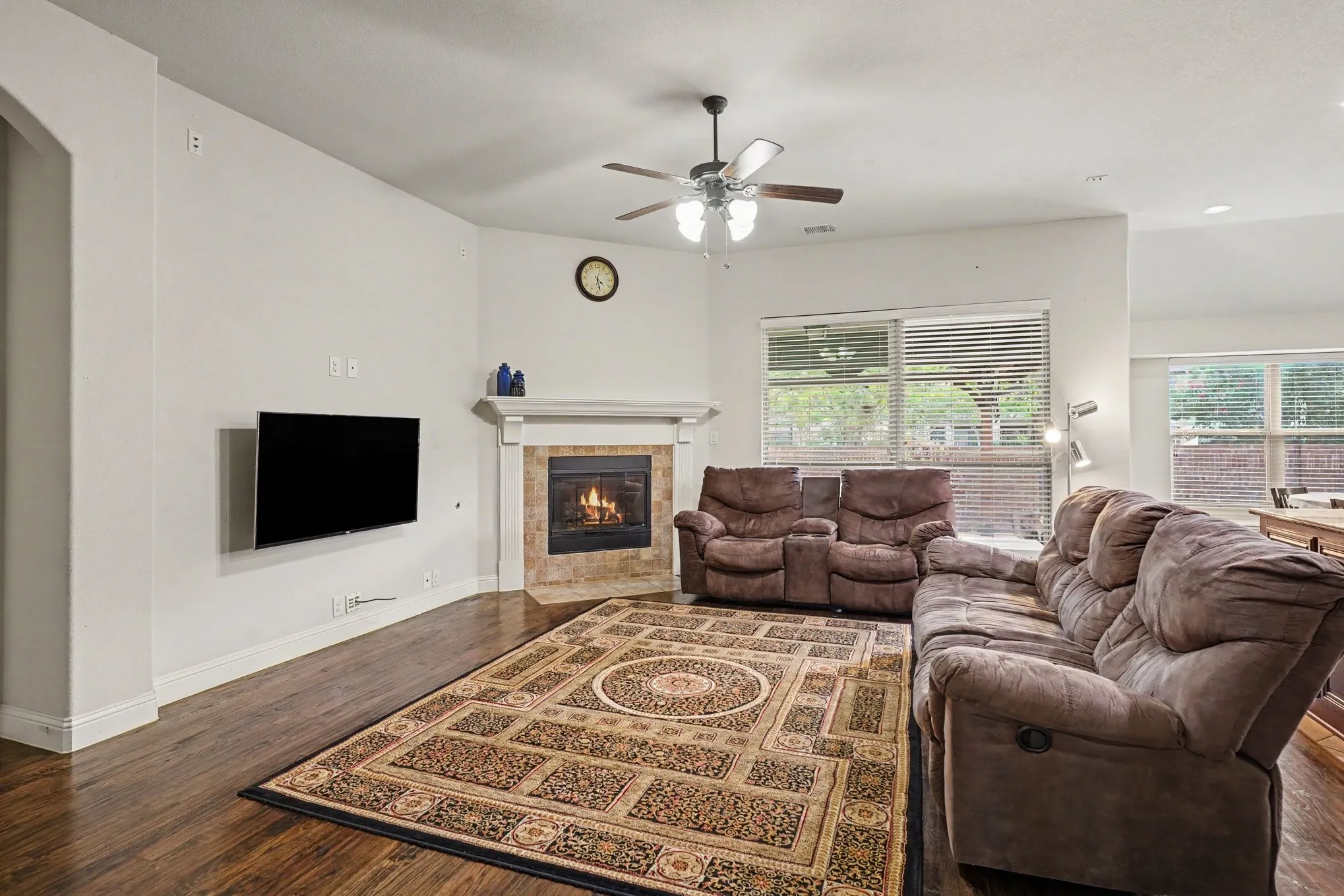 Wood-finish flooring, corner fireplace with a mantel, ceiling fan with integrated lighting, multiple windows with blinds, and white walls