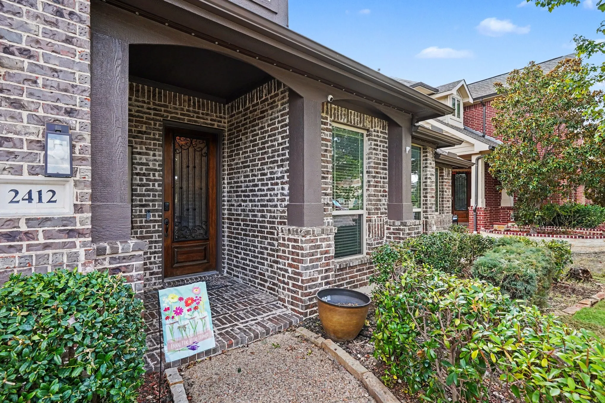 Brick exterior facade with a covered entry, dark wood-finish door, exterior wall lantern, and mature shrubbery