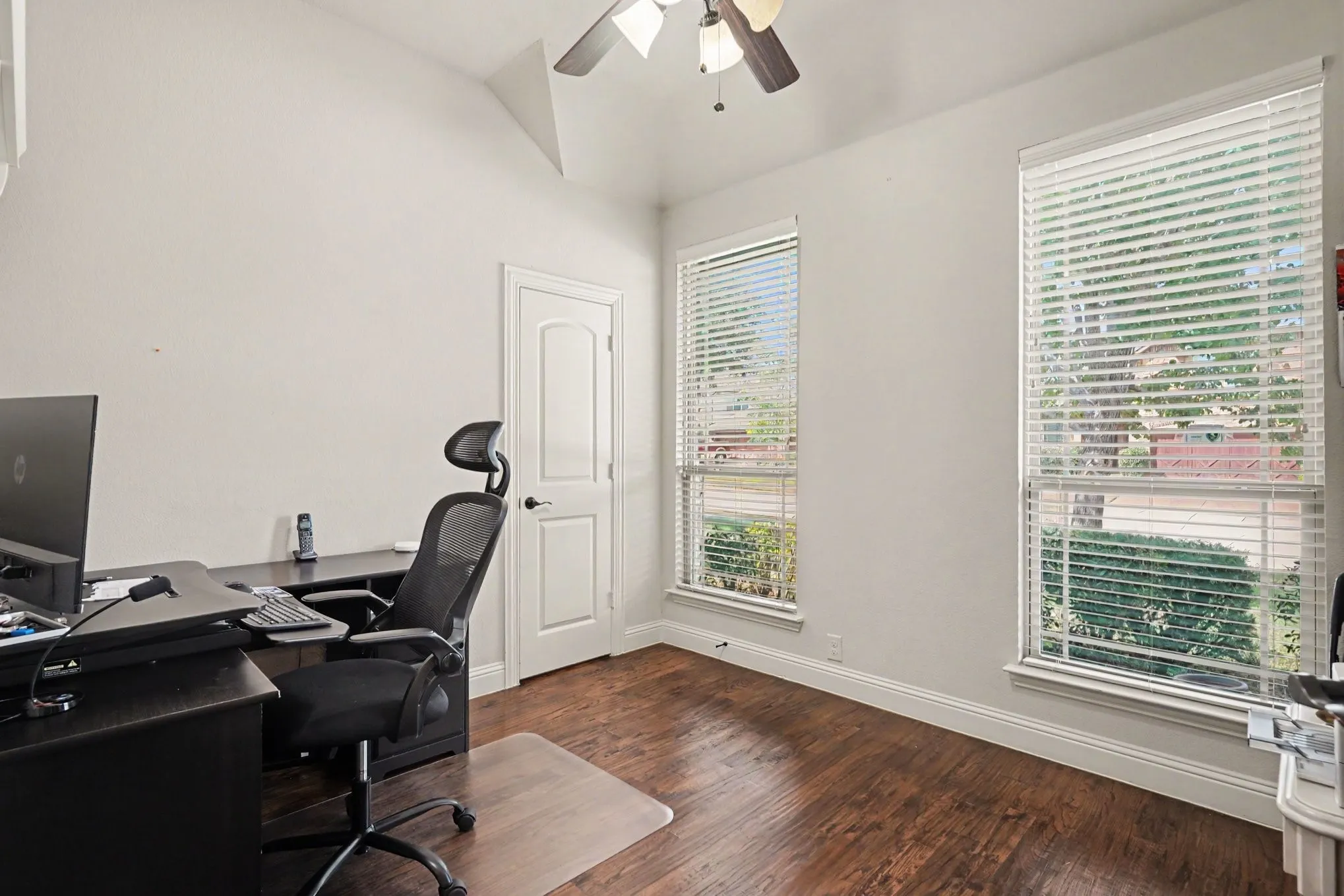 Vaulted ceiling with a ceiling fan, two windows with blinds, wood-finish flooring, and a white paneled door