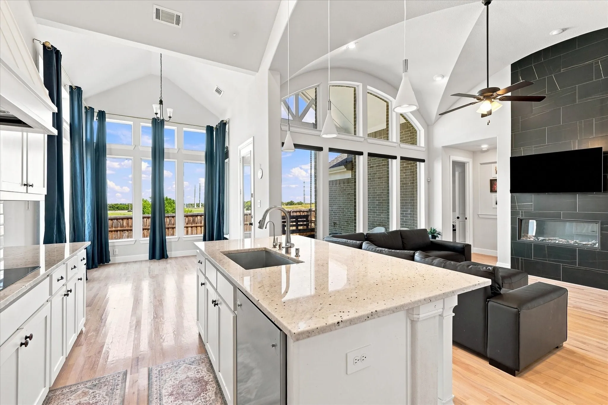 Open-concept living area with vaulted ceilings, a central kitchen island featuring a stainless steel sink, large window expanses, and a fireplace with a dark tile surround
