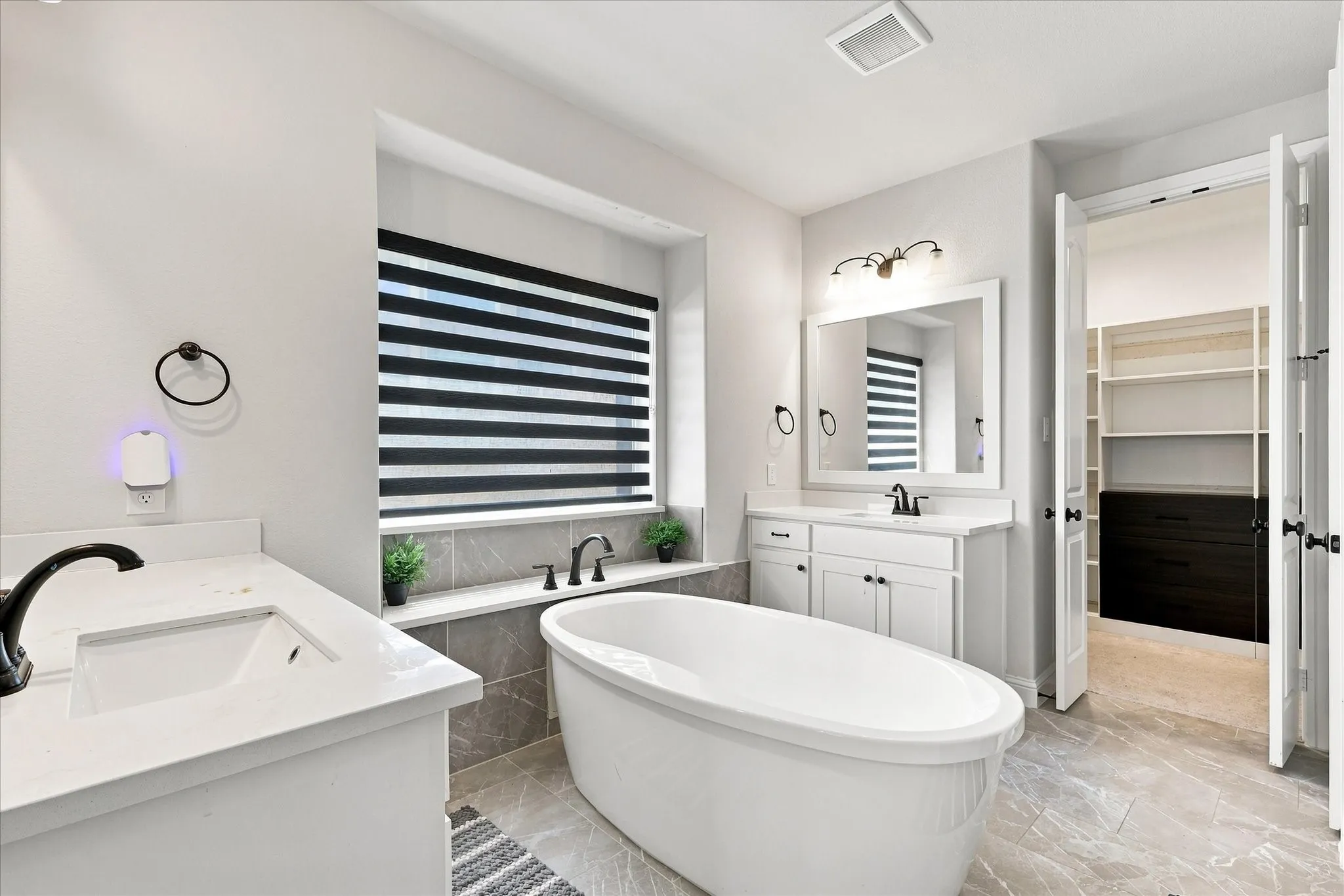 Freestanding soaking tub, dual vanities with white countertops, light gray marble-look tile flooring, and a walk-in closet with built-in shelving