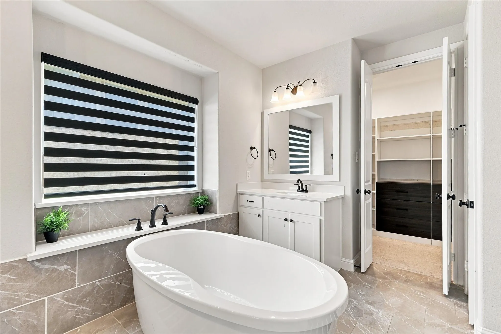 Freestanding soaking tub with a matte black faucet, single vanity with white cabinetry, framed mirror, and a built-in closet system
