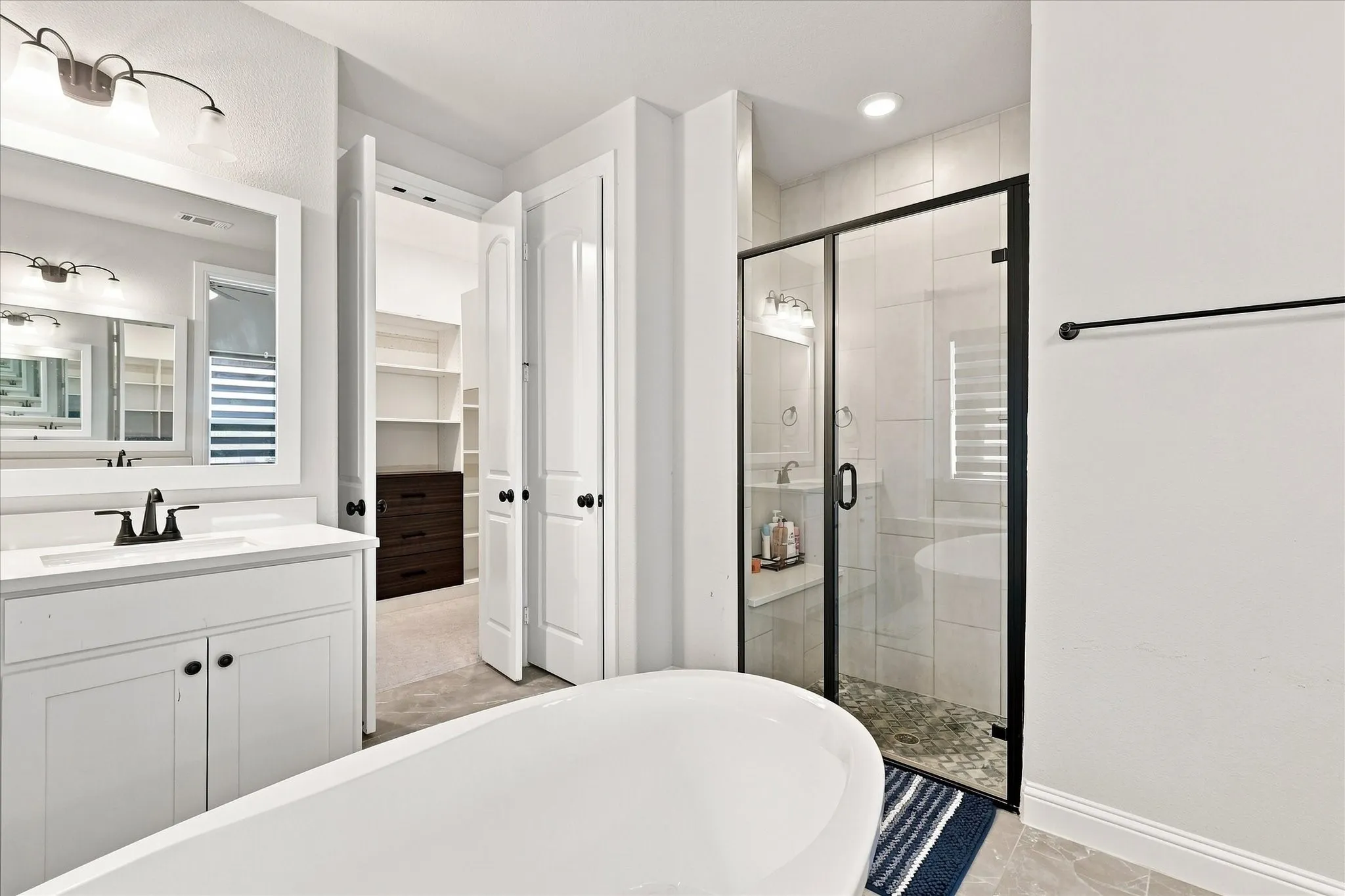 Freestanding soaking tub, walk-in shower with glass enclosure, white vanity with integrated sink, walk-in closet with built-in shelving, and tile flooring