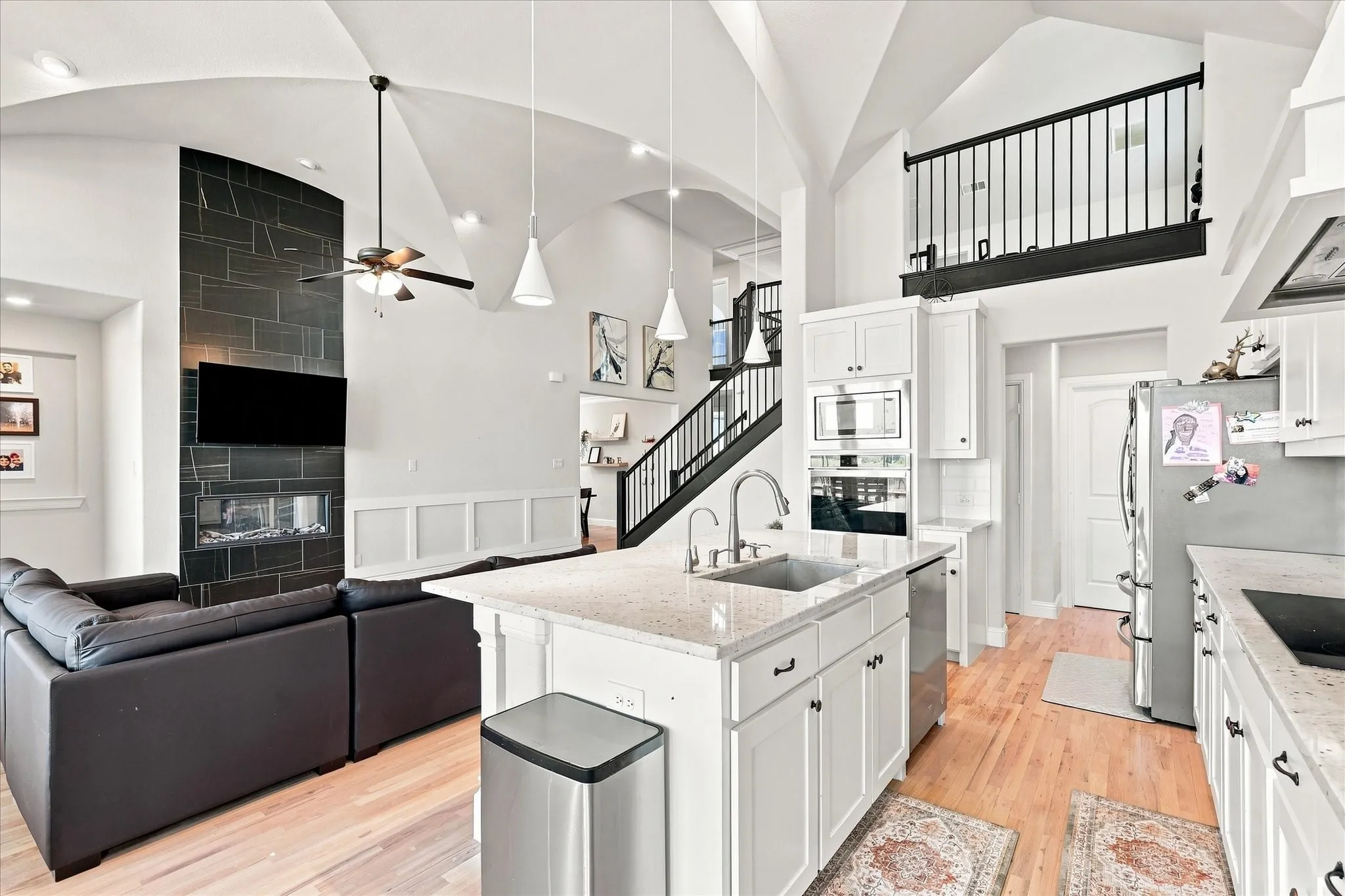 Vaulted ceiling with arch details, wood-finish flooring, kitchen island with integrated sink, built-in oven and microwave, and a fireplace with a dark tile surround