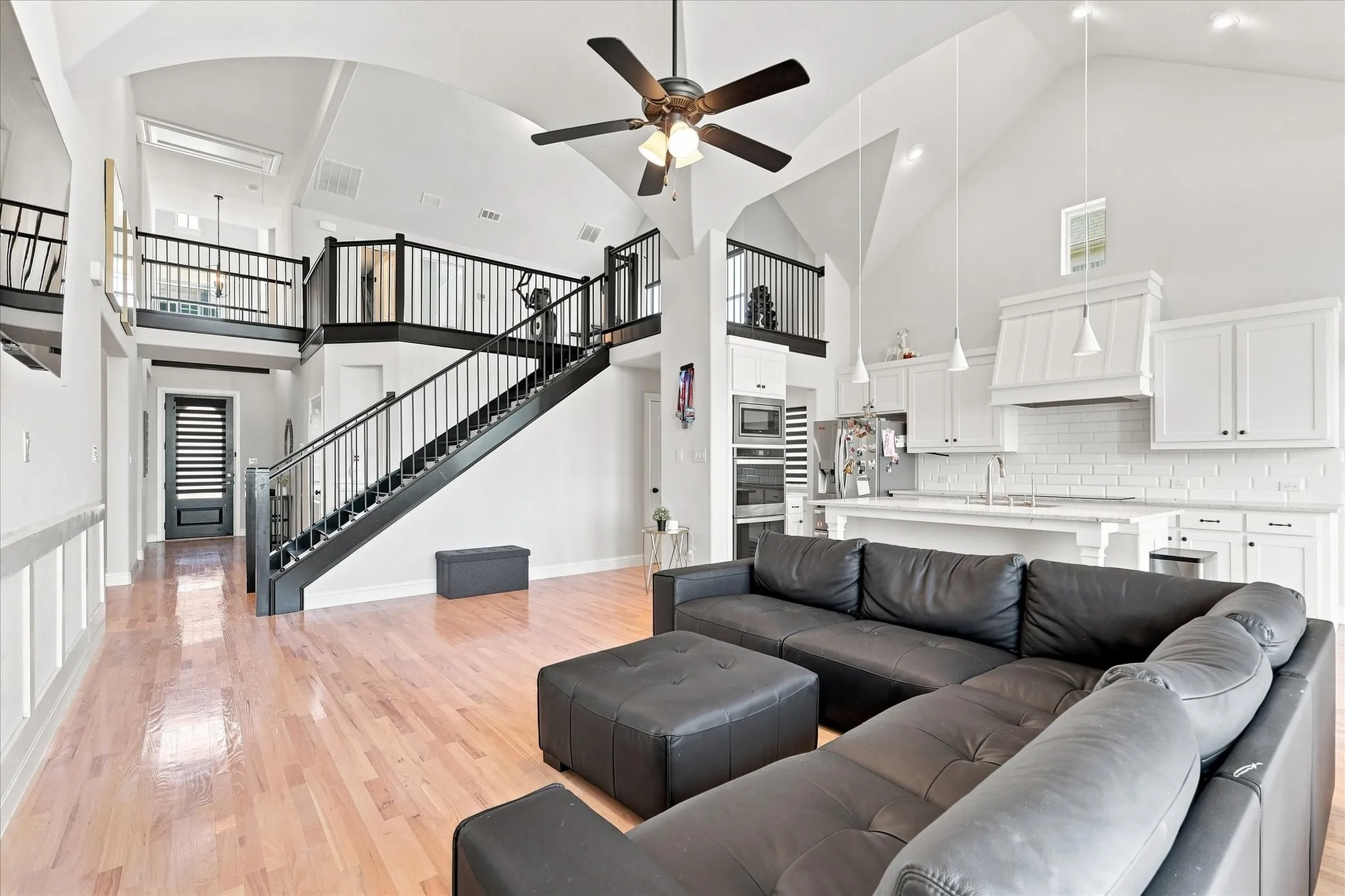 Vaulted ceilings, wood-finish flooring, staircase with metal balusters, kitchen island with built-in sink, and white kitchen cabinetry