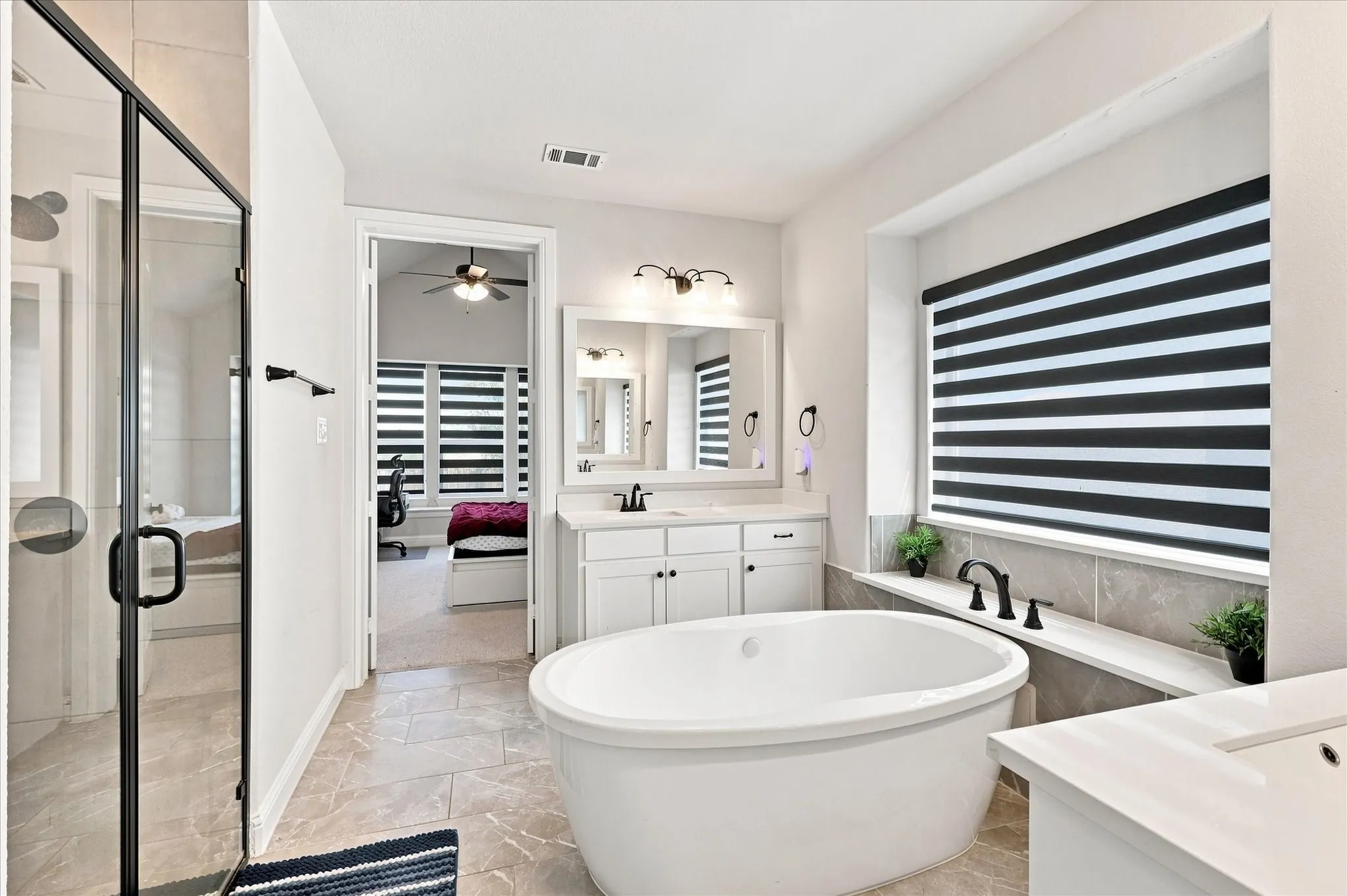 Freestanding bathtub, dual vanity with white cabinetry, glass-enclosed shower with black trim, black-and-white striped window treatment, and large format tile flooring