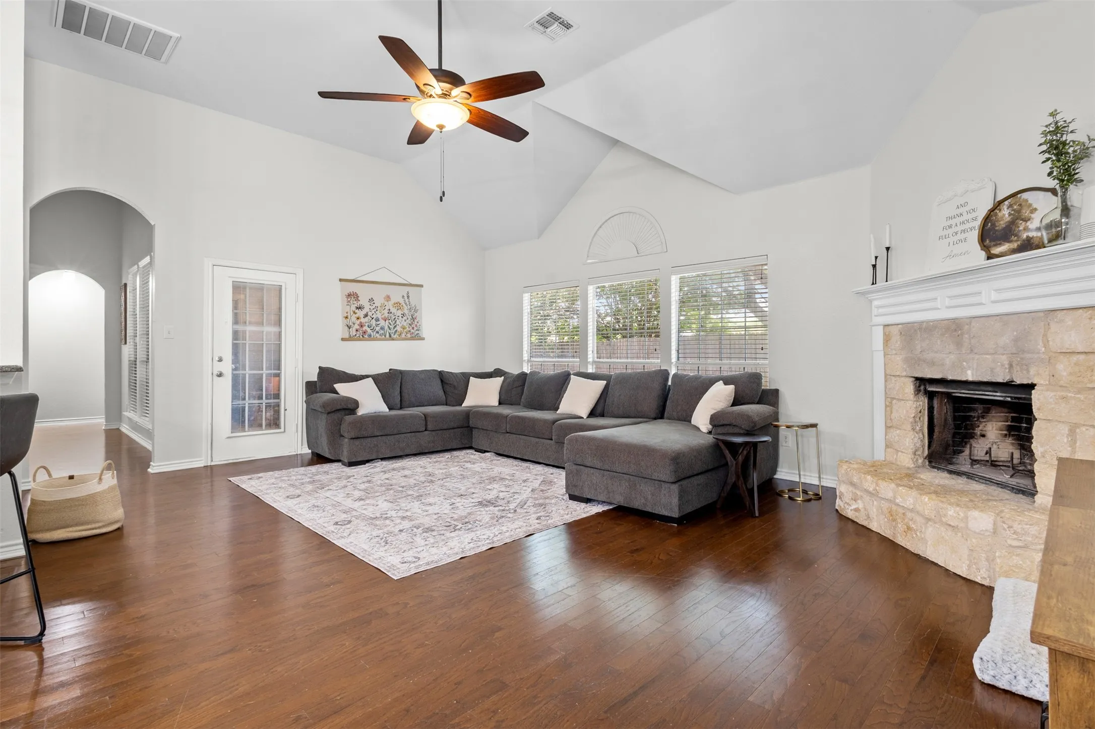 Living area featuring vaulted ceilings, wood-finish flooring, a stone-faced fireplace with a mantel, a ceiling fan, and large windows