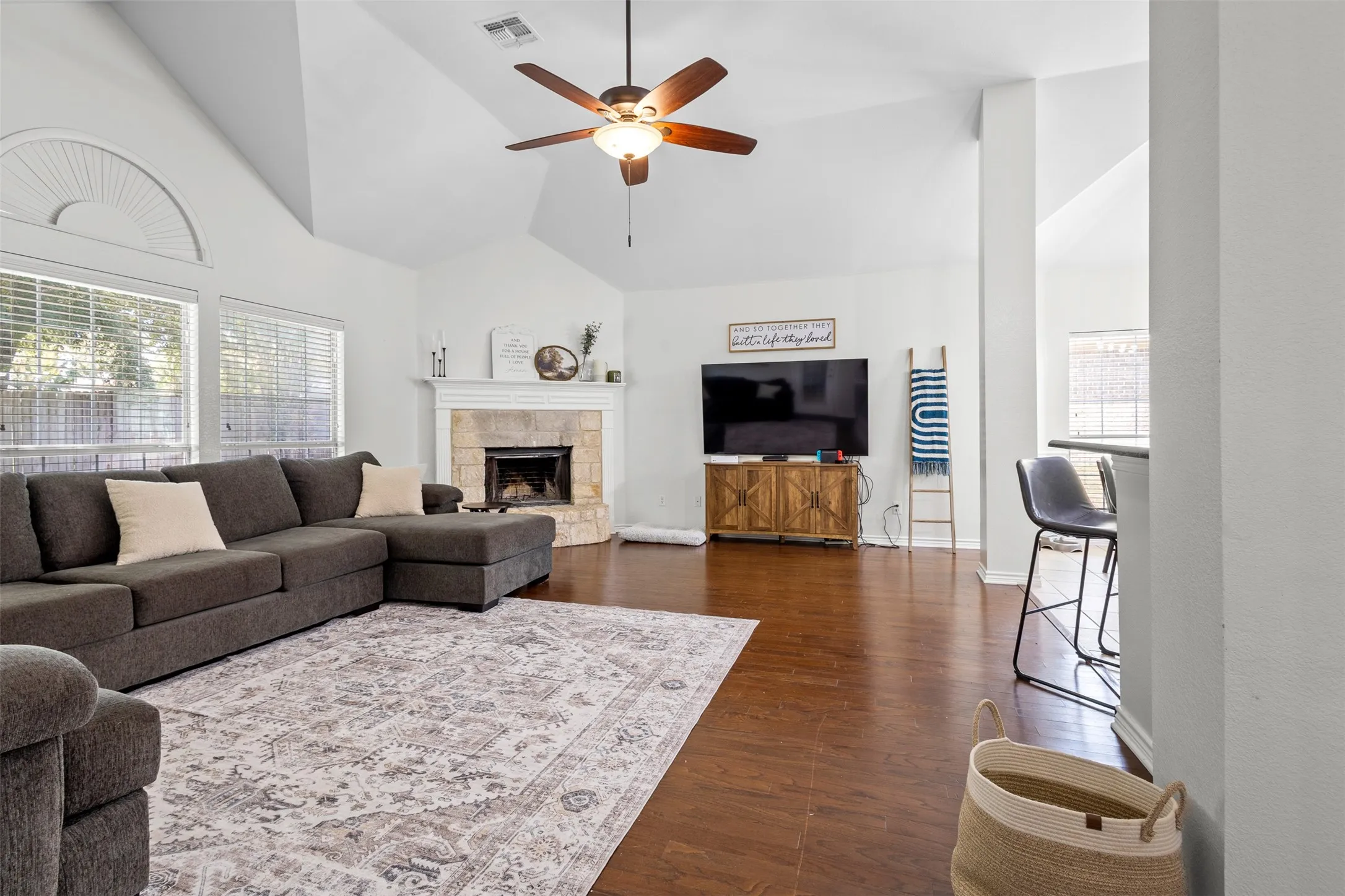 Vaulted ceiling, wood-finish flooring, stone-surround fireplace, ceiling fan, and large windows with arched transom