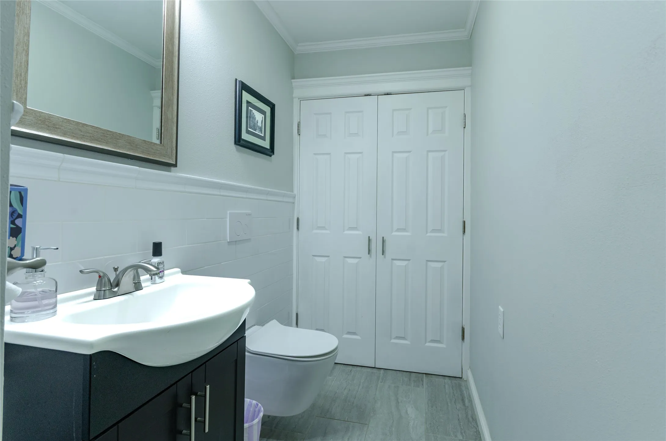 Wall-mounted toilet, vanity with integrated basin, tiled wainscoting, and crown molding