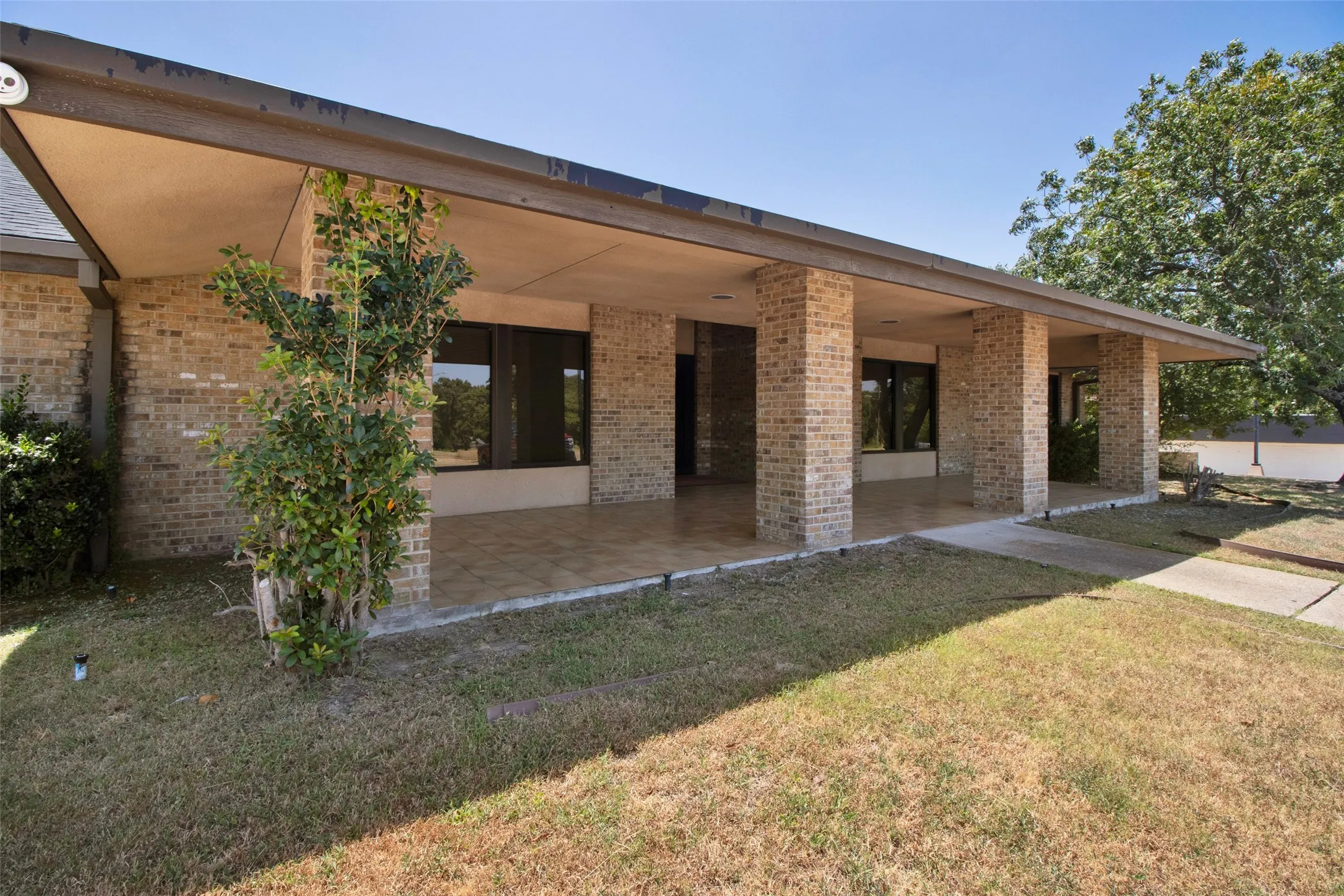Brick exterior with an expansive covered patio, featuring multiple support columns, large windows, and a paved walkway