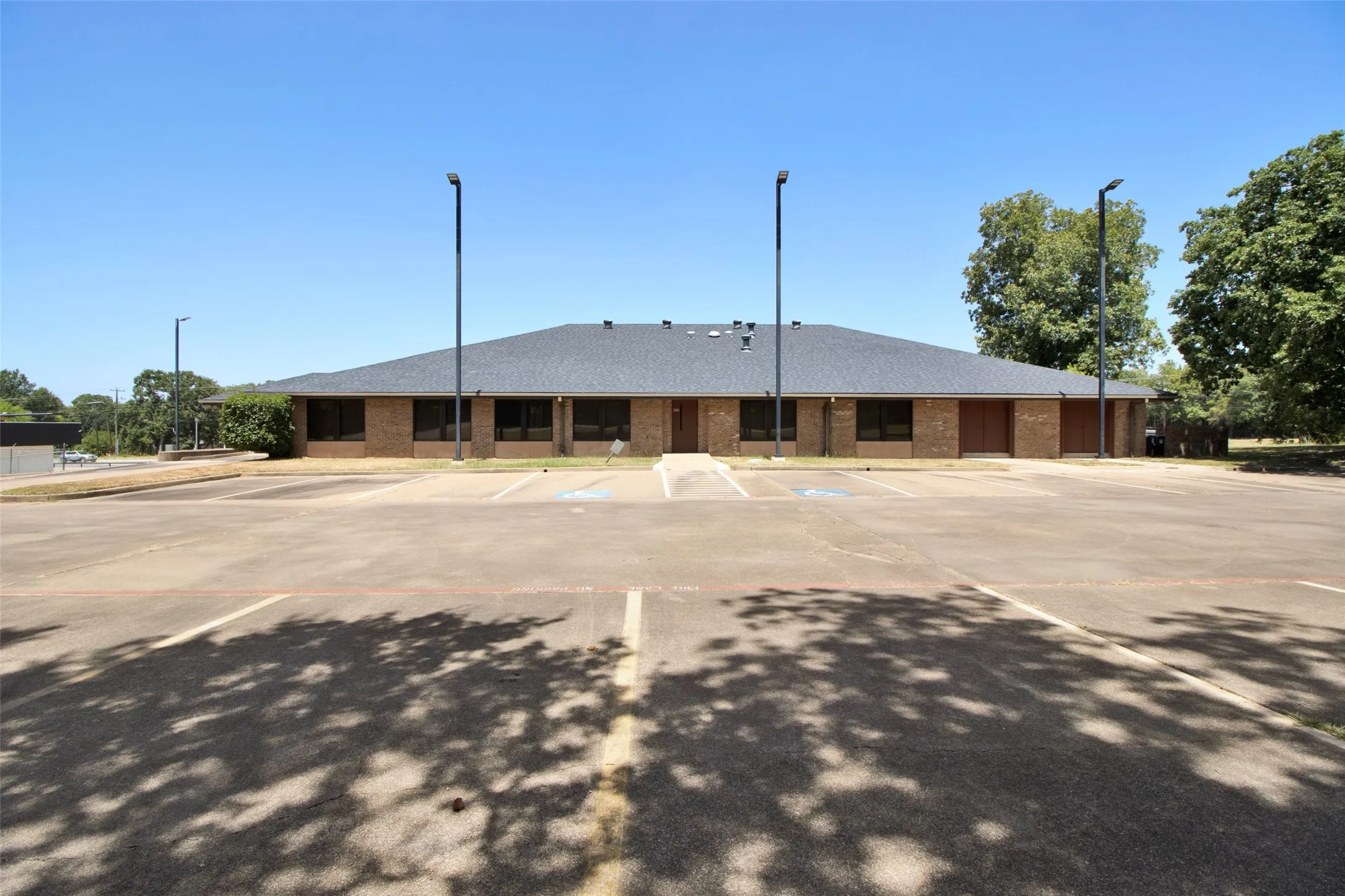 Brick exterior with a dark shingle roof, expansive paved parking lot, and multiple exterior lighting fixtures