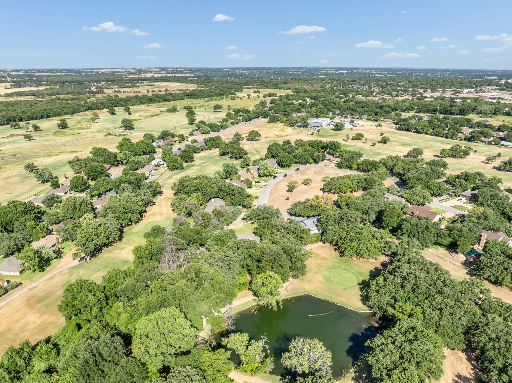 Aerial panorama featuring a golf course, scattered residences, a natural pond, and extensive tree coverage