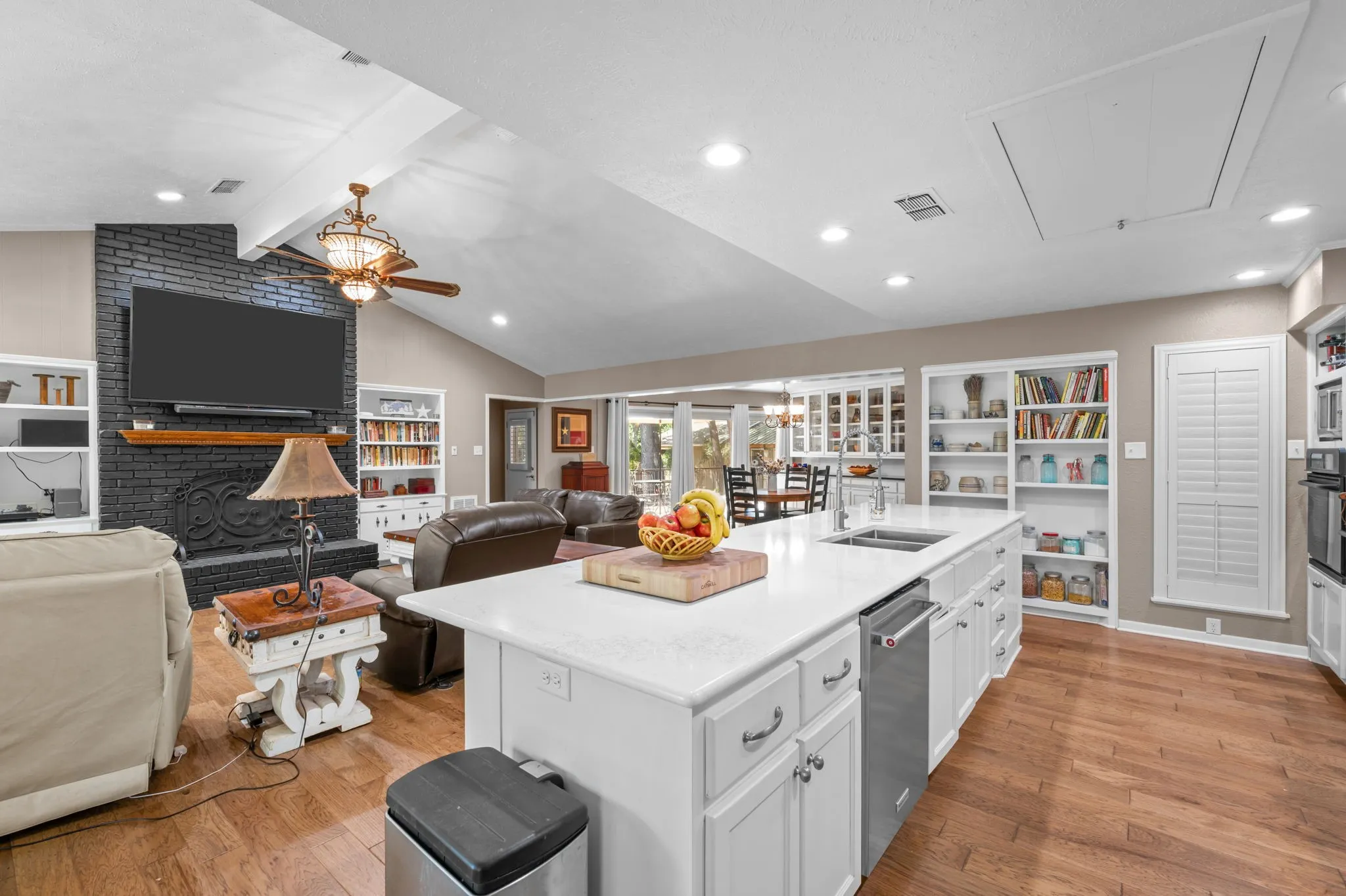 Open-concept living area with vaulted ceilings, a central kitchen island with integrated sink, wood-finish flooring, and a black brick fireplace with a wood mantel