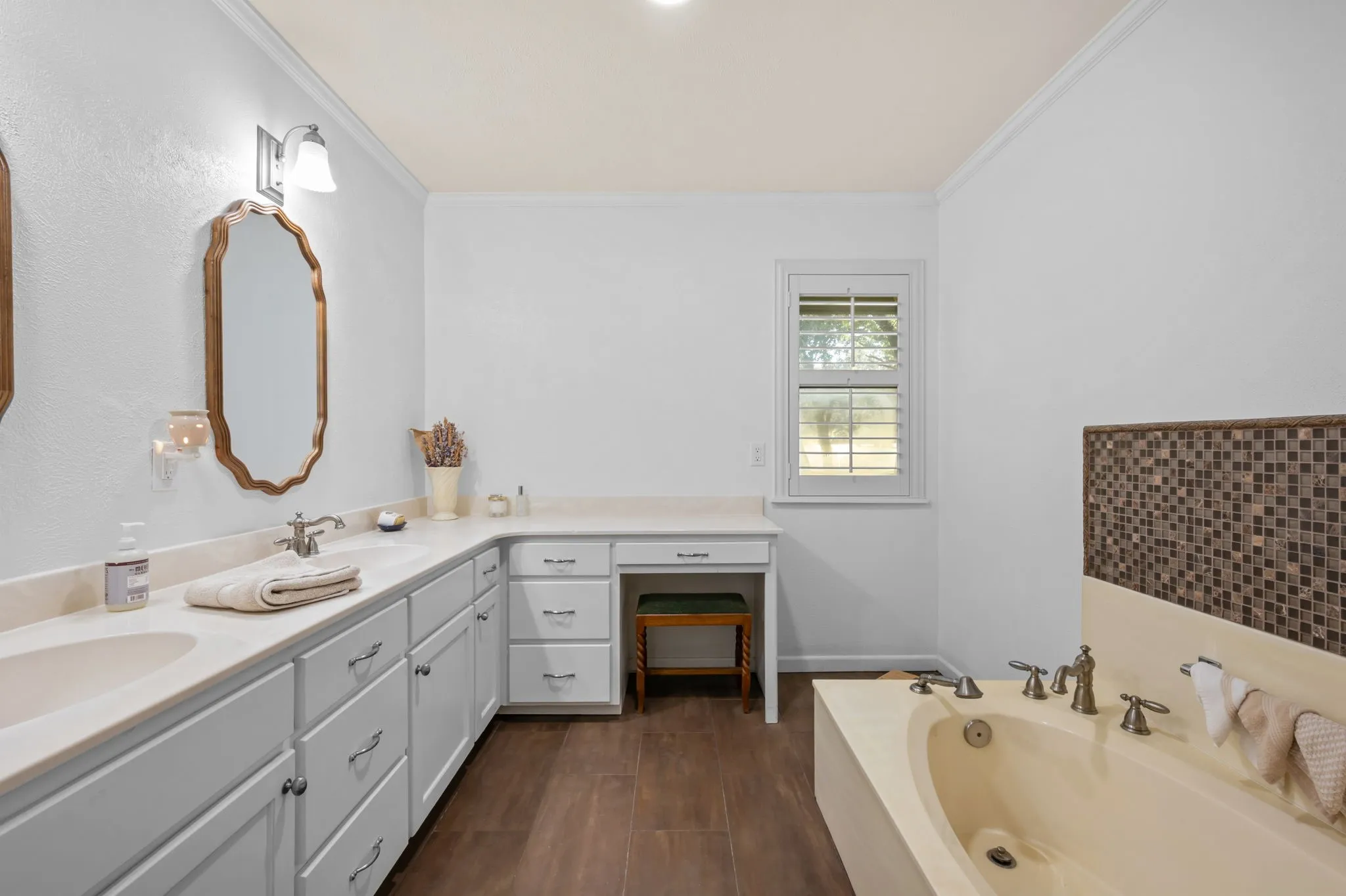 Dual vanity with under-mount sinks and light-toned countertops, extensive white cabinetry with brushed nickel hardware, built-in vanity desk, wood-finish flooring, and a built-in soaking tub with mosaic tile surround