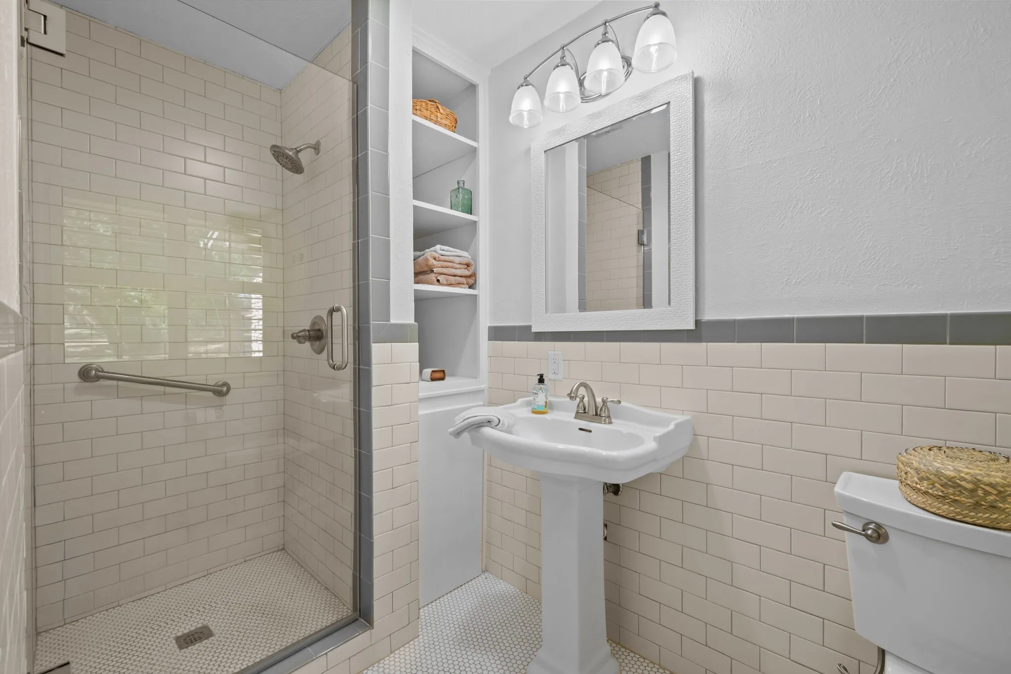 Glass-enclosed shower with subway tile surround, built-in shelving, pedestal sink, and vanity mirror with sconce lighting