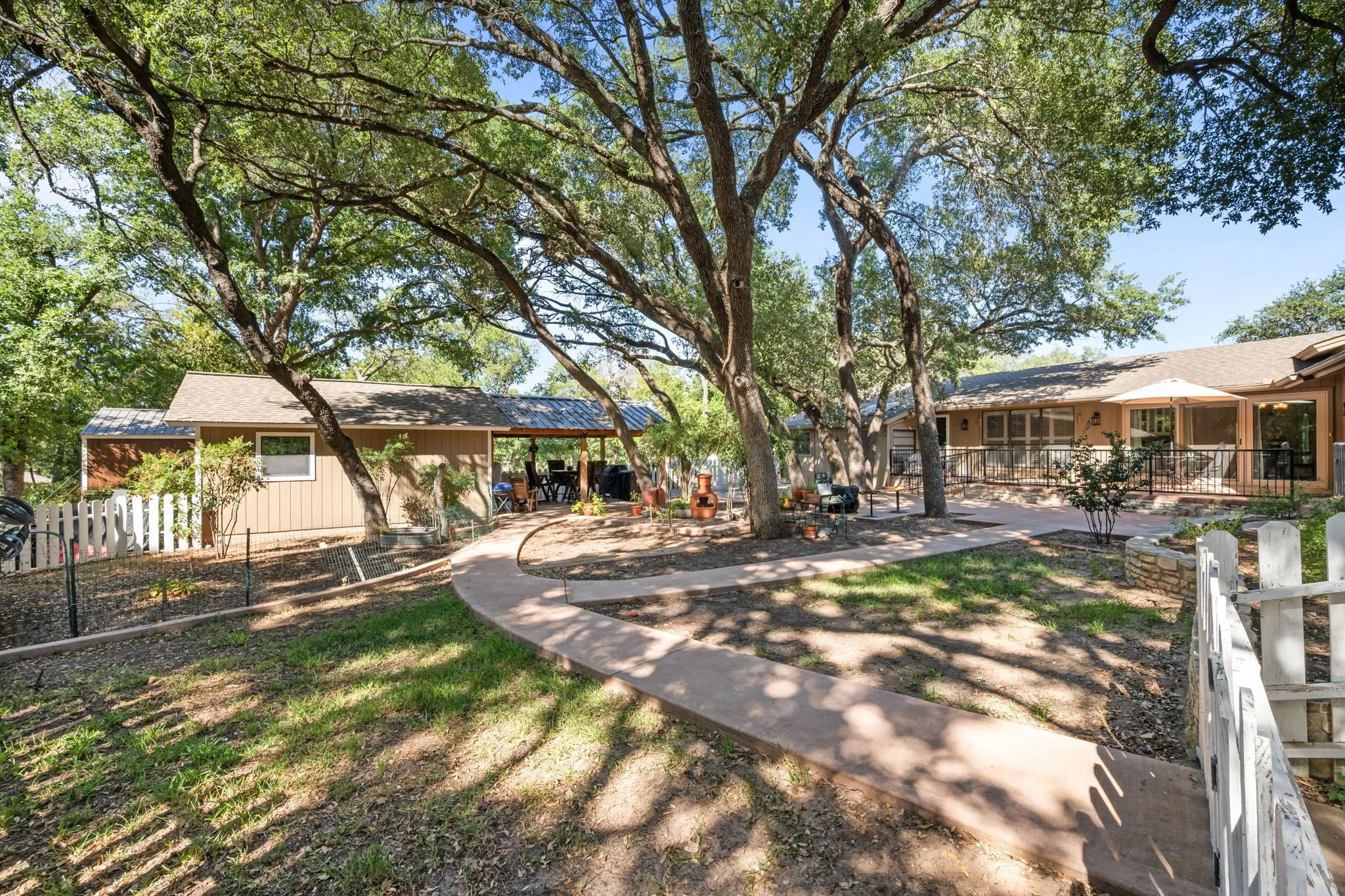 Expansive outdoor living area featuring multiple mature trees, a curved concrete pathway, a wooden picket fence, a covered patio structure, and an additional building