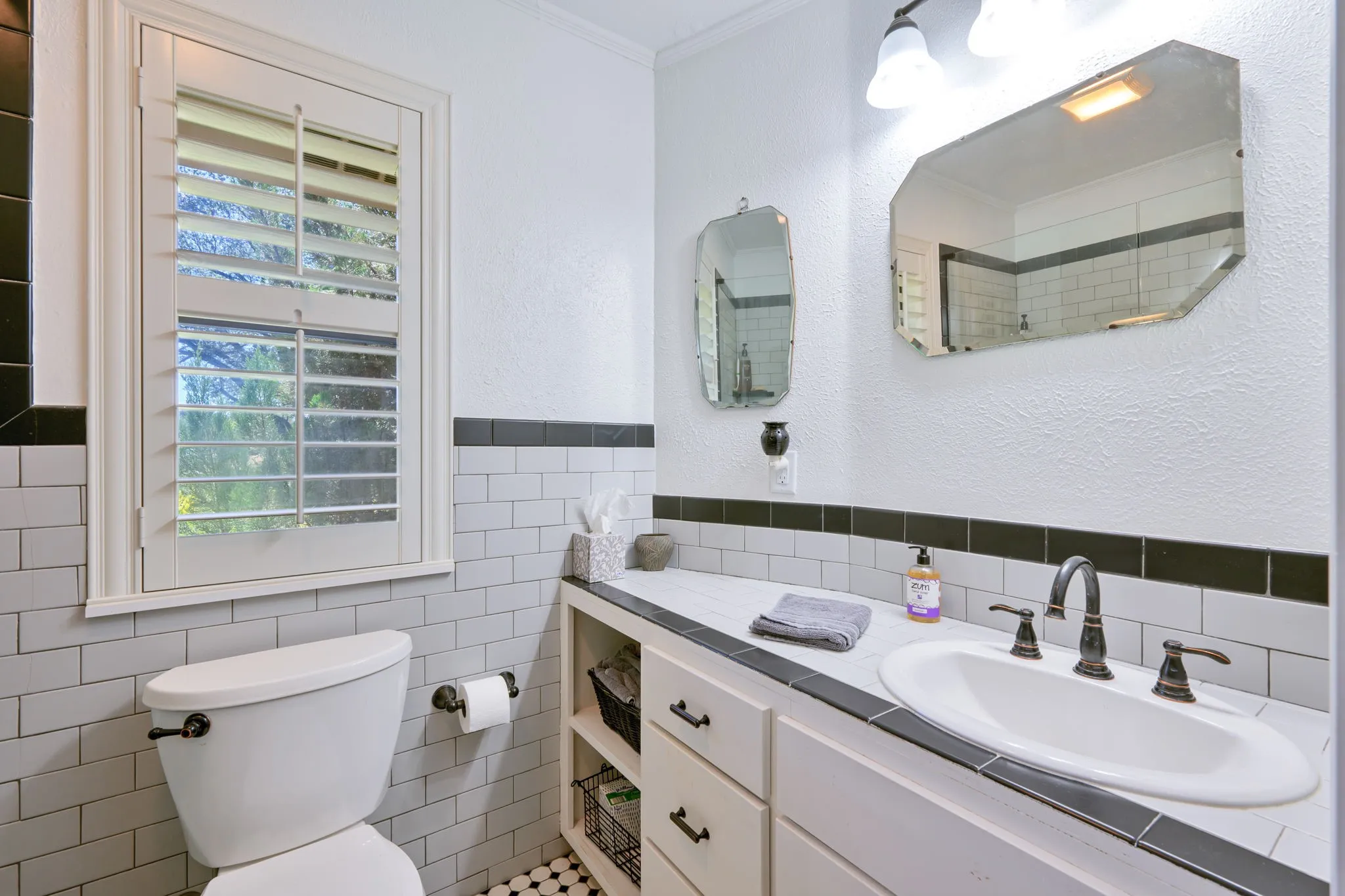 White subway tile, a vanity with integrated sink, black and white floor tile, a window with plantation shutters, and a wall-mounted light fixture