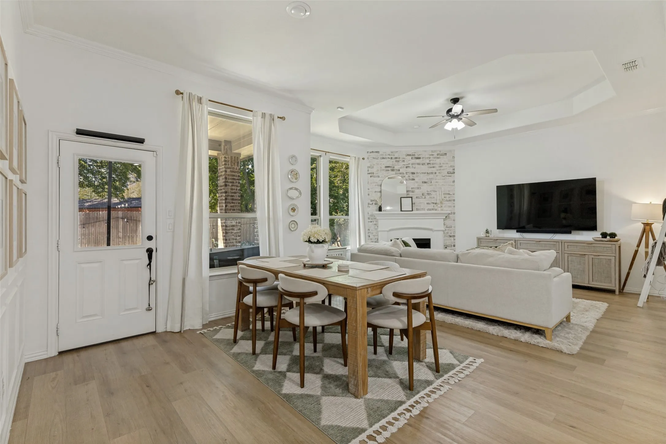 Open-concept living space with wood-finish flooring, a tray ceiling with a ceiling fan, and a fireplace featuring a white brick-finish surround