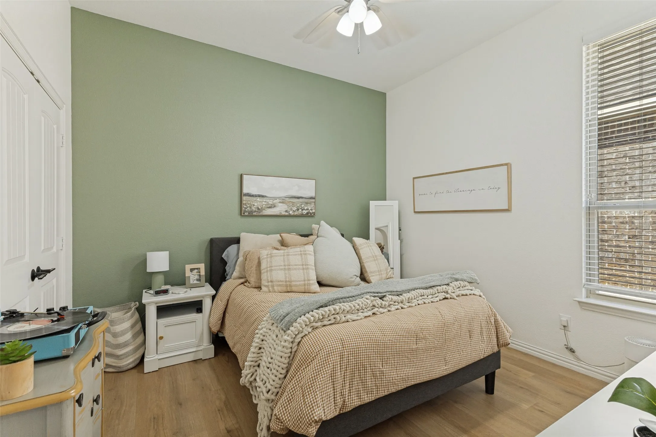 Wood-finish flooring, accent wall, ceiling fan, window with blinds, and white door with black hardware