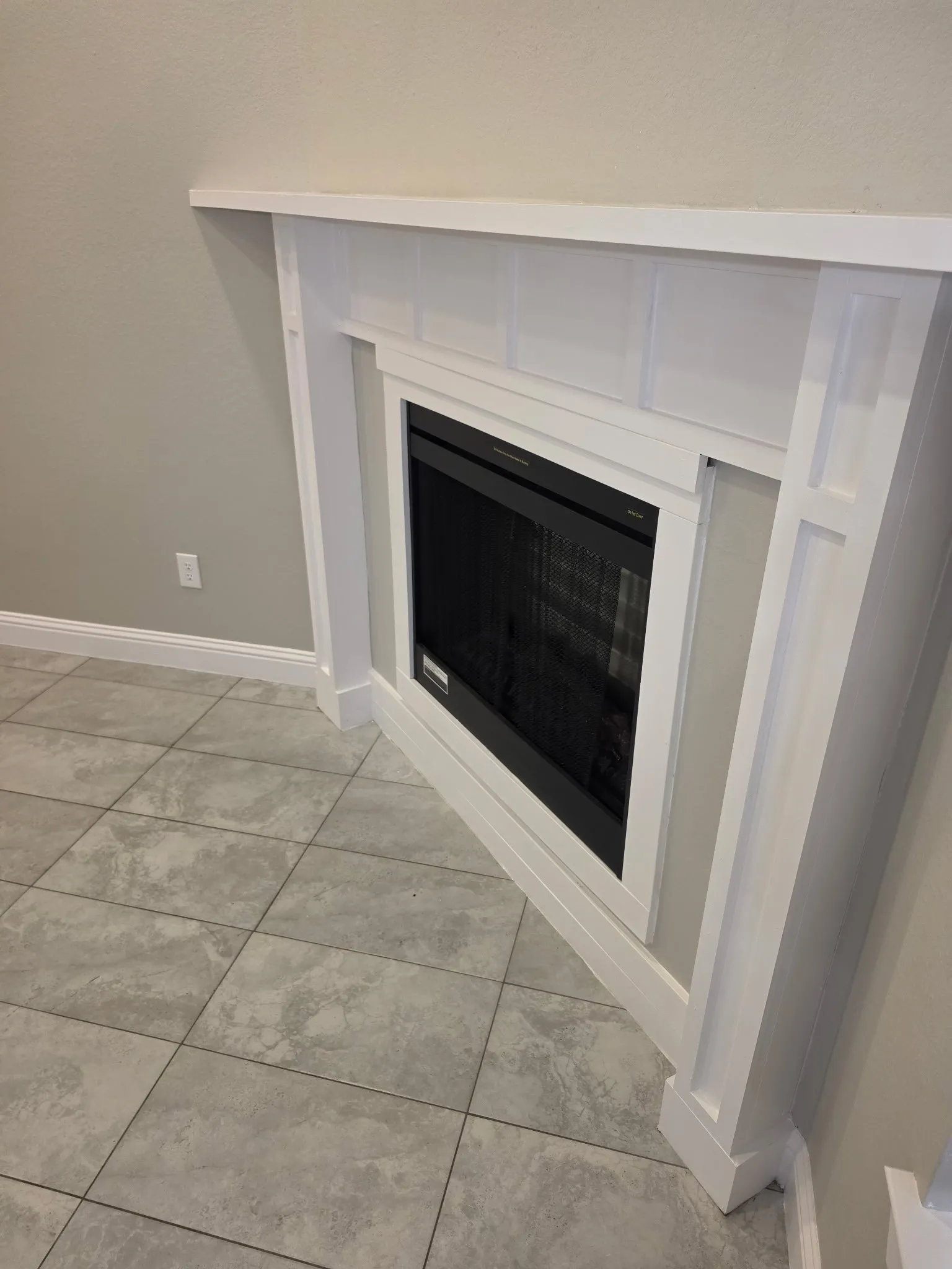 Built-in corner electric fireplace with white mantel and surround, set against light gray walls and large format tile flooring