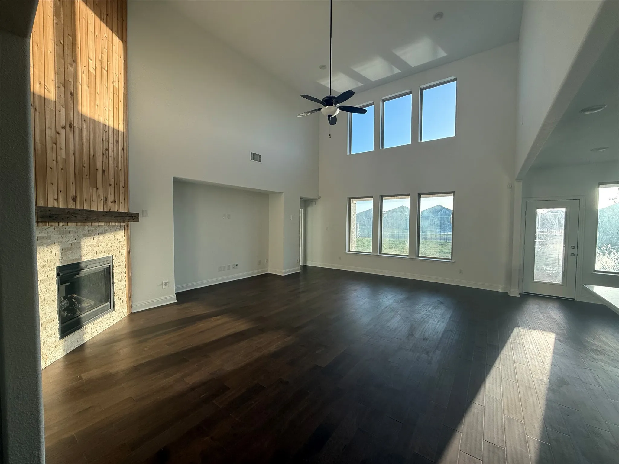 Vaulted ceiling with a ceiling fan, fireplace with a stone surround and wood-finish accent wall, dark wood-finish flooring, and multiple windows