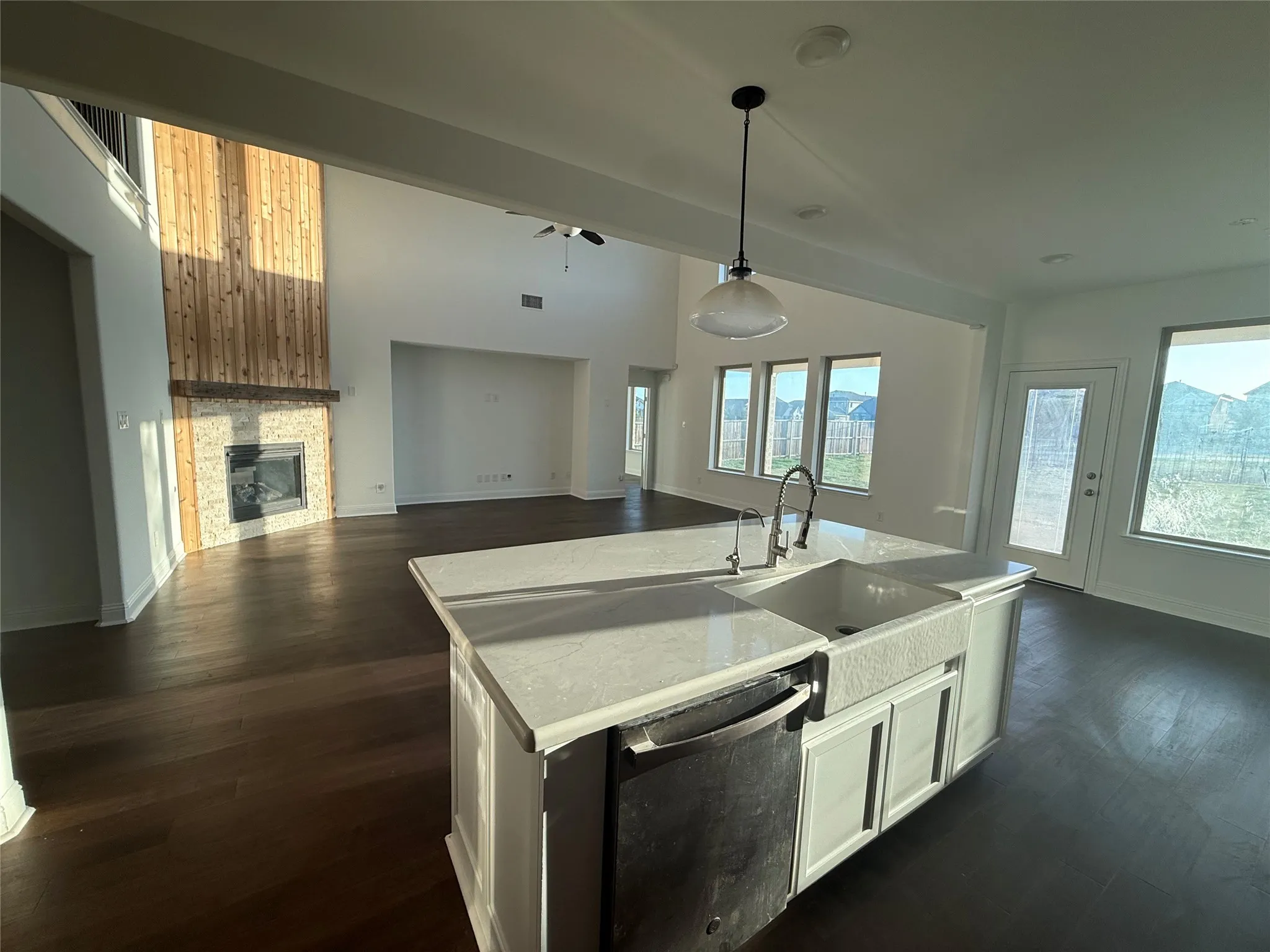 Expansive open-concept living area with dark wood-finish flooring, a white kitchen island featuring a farmhouse sink, and a fireplace with a stone surround and wood paneling