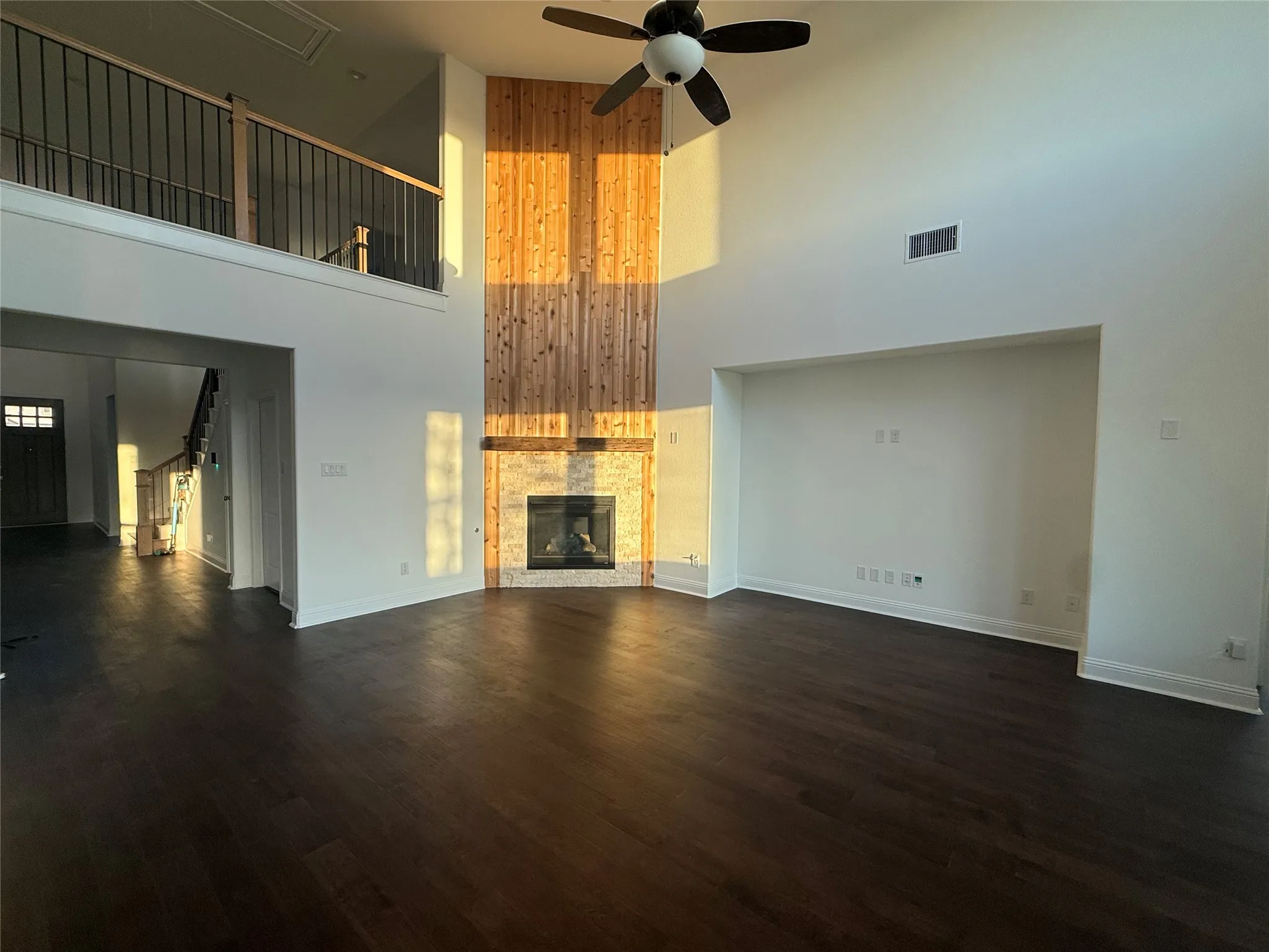 Expansive living area featuring soaring ceilings, a wood-finish flooring, a contemporary ceiling fan, and a fireplace with a stone surround and wood paneling