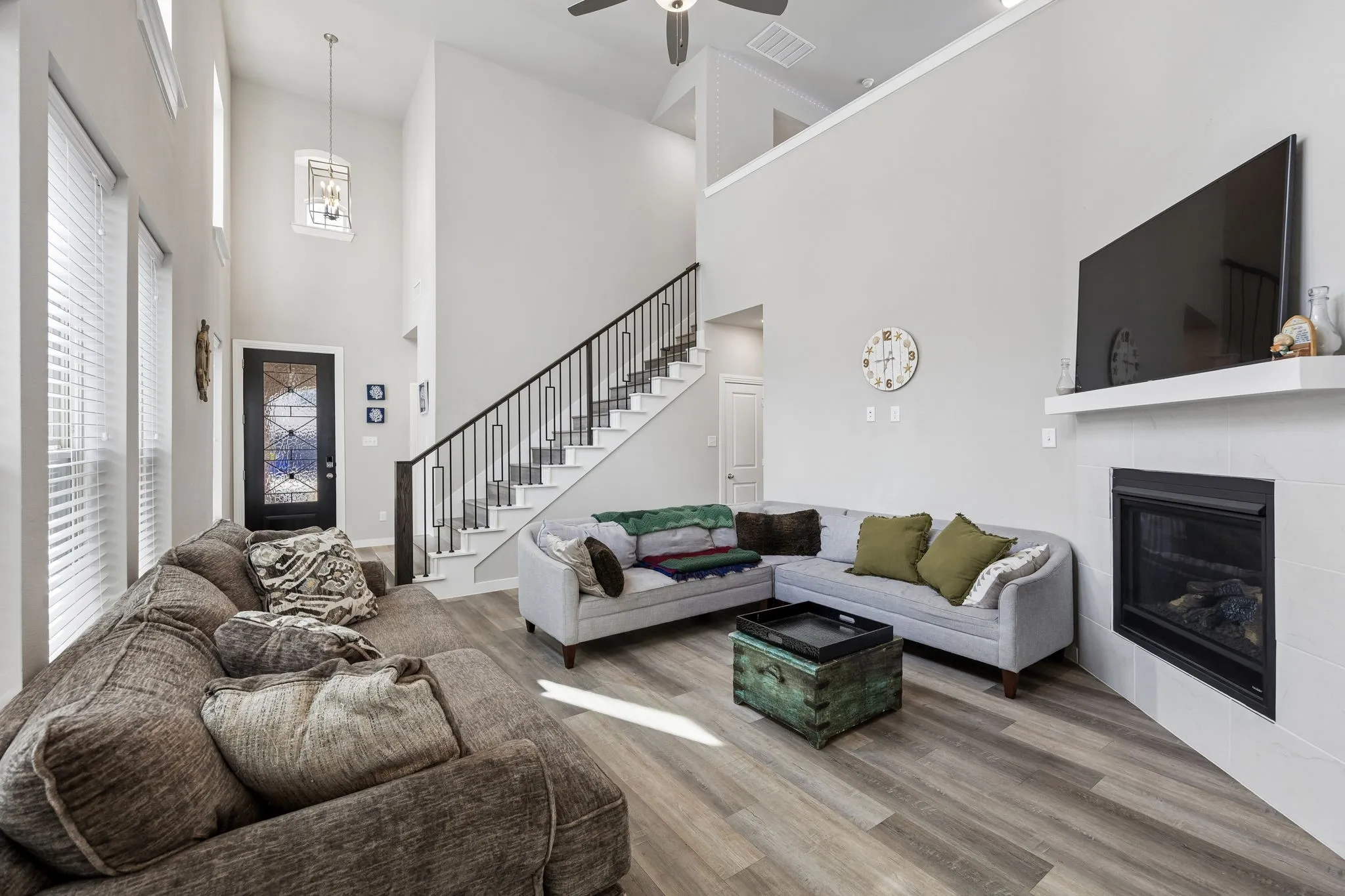 Two-story ceiling, wood-finish flooring, white tile fireplace with mantle, integrated fireplace, and a staircase with wrought iron balusters